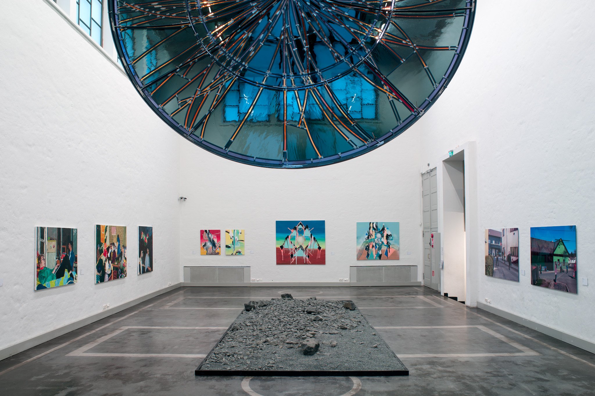 An art gallery with white walls displaying colorful paintings. A large, modern sculpture made of rocks sits in the center on a rectangular black frame. A glass ceiling with a blue and orange metal structure is above, and a doorway is visible to the right.