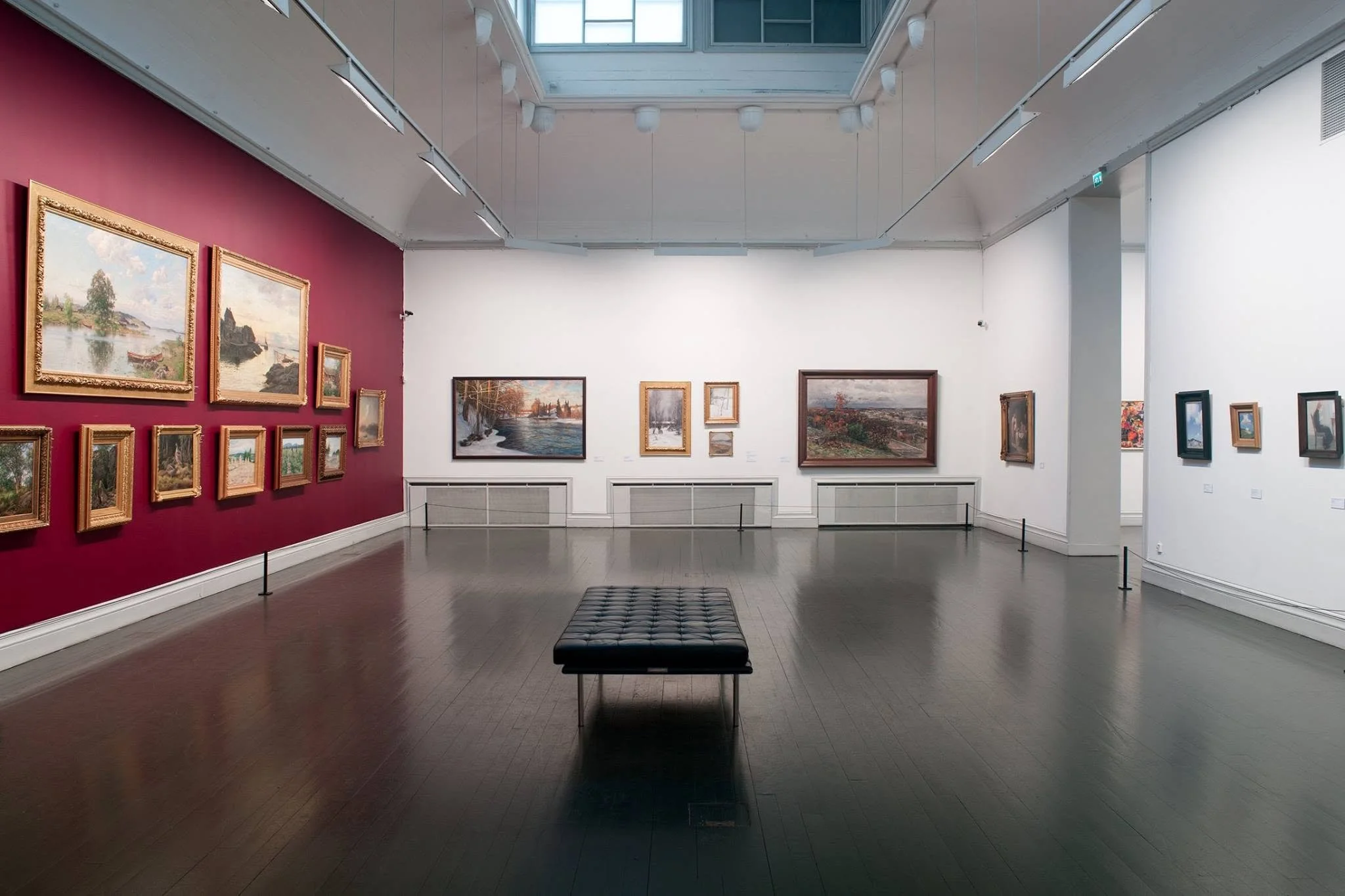 An art gallery with paintings of landscapes hung on white and burgundy walls. There is a black bench in the center of the room, and a skylight overhead allowing natural light to illuminate the artwork.