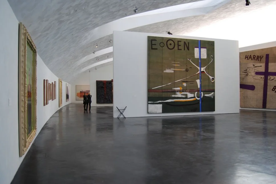 Interior of an art gallery with abstract paintings and sculptures on white walls and a curved ceiling, with a few visitors viewing the artworks.