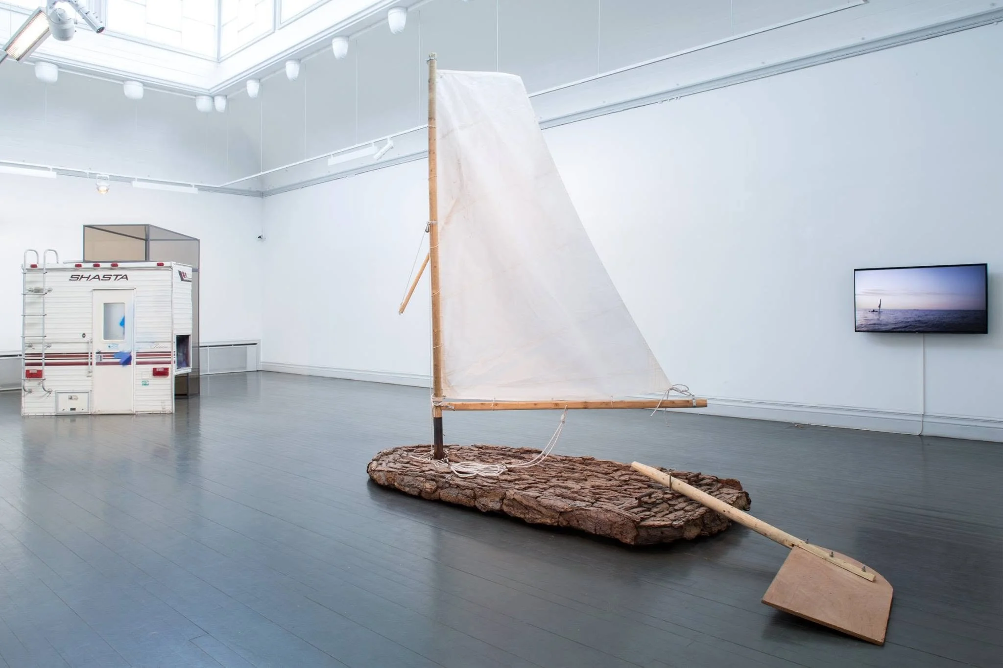 Art installation featuring a small sailboat model made of wood and fabric, mounted on a large disk of tree bark, displayed in a bright gallery space with a television showing a sailboat on water and a Tuff Shed in the background.