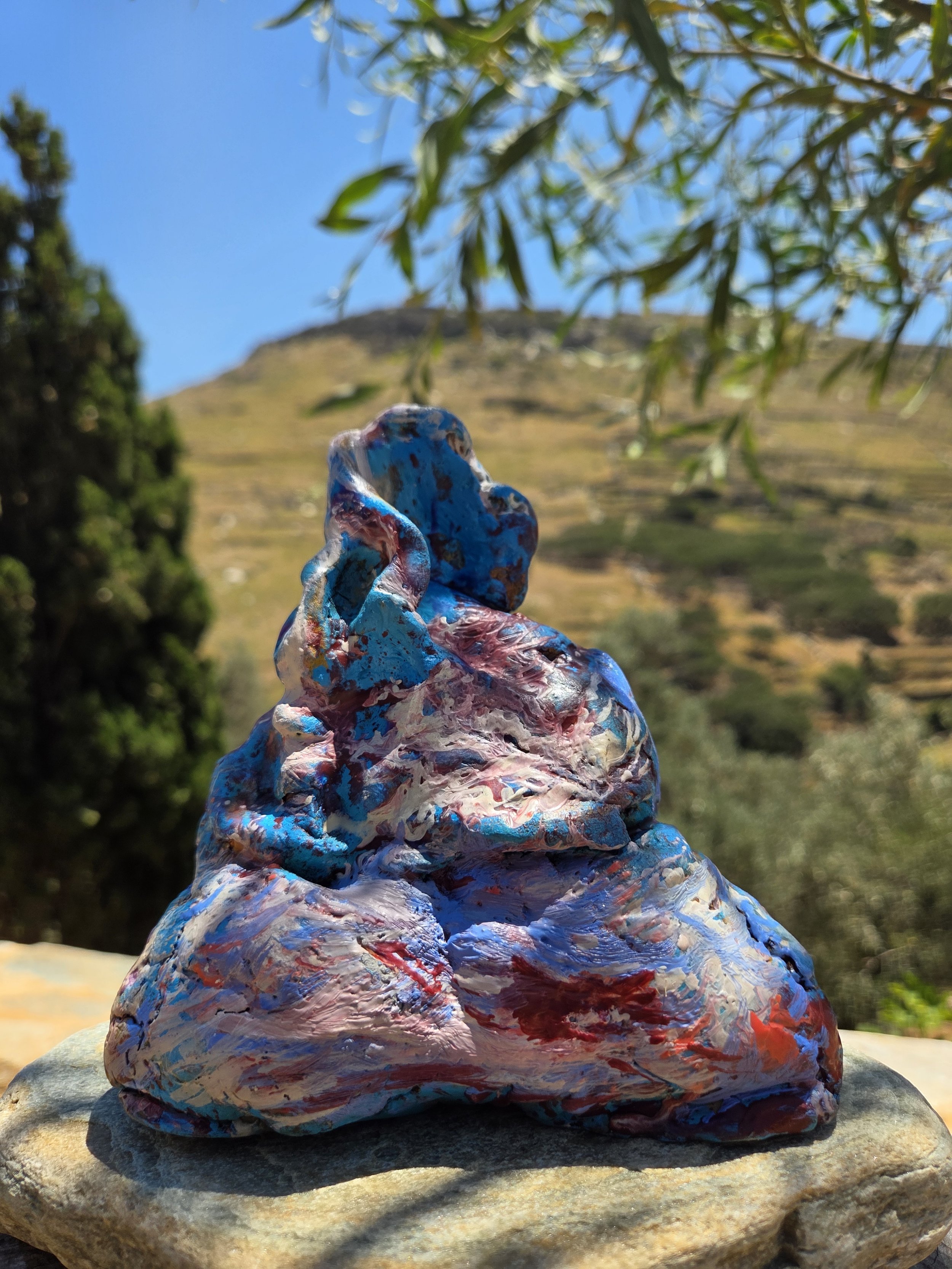 Painted art work rock with colorful swirls and textures, sitting on a flat stone surface outdoors, with a blurred mountain, blue sky, and tree branches in the background. Abstract sulpture by Finnish artist Satu Metsola