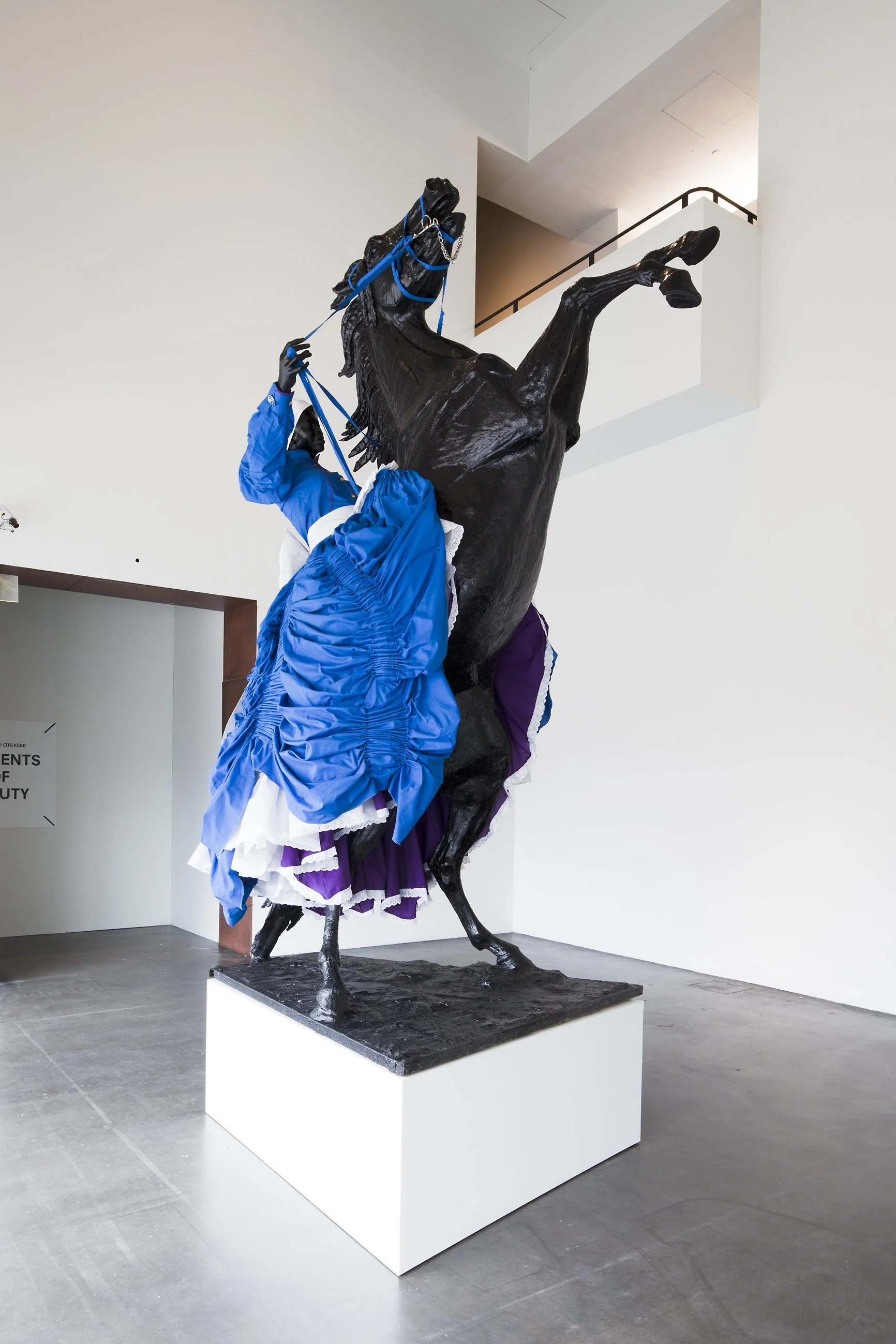 A sculpture of a rearing horse, painted black, adorned with a purple and white ruffled skirt and a woman in a blue jacket adjusting a harness on the horse's head.