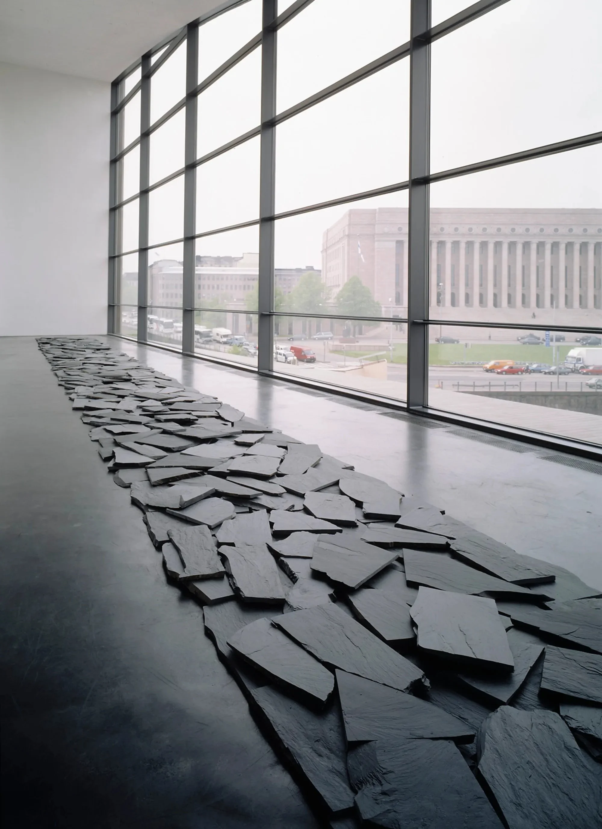 An art installation with a large glass window overlooking a city street, featuring a scattered arrangement of white, irregularly shaped slabs on the floor.