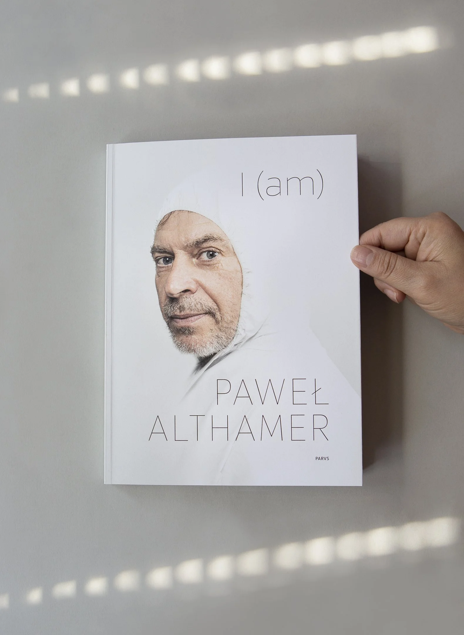 A person holding a white book titled "I (am)" by Pawel Althamer. The cover features a partial face of an older man with gray hair, a beard, blue eyes, and a serious expression.