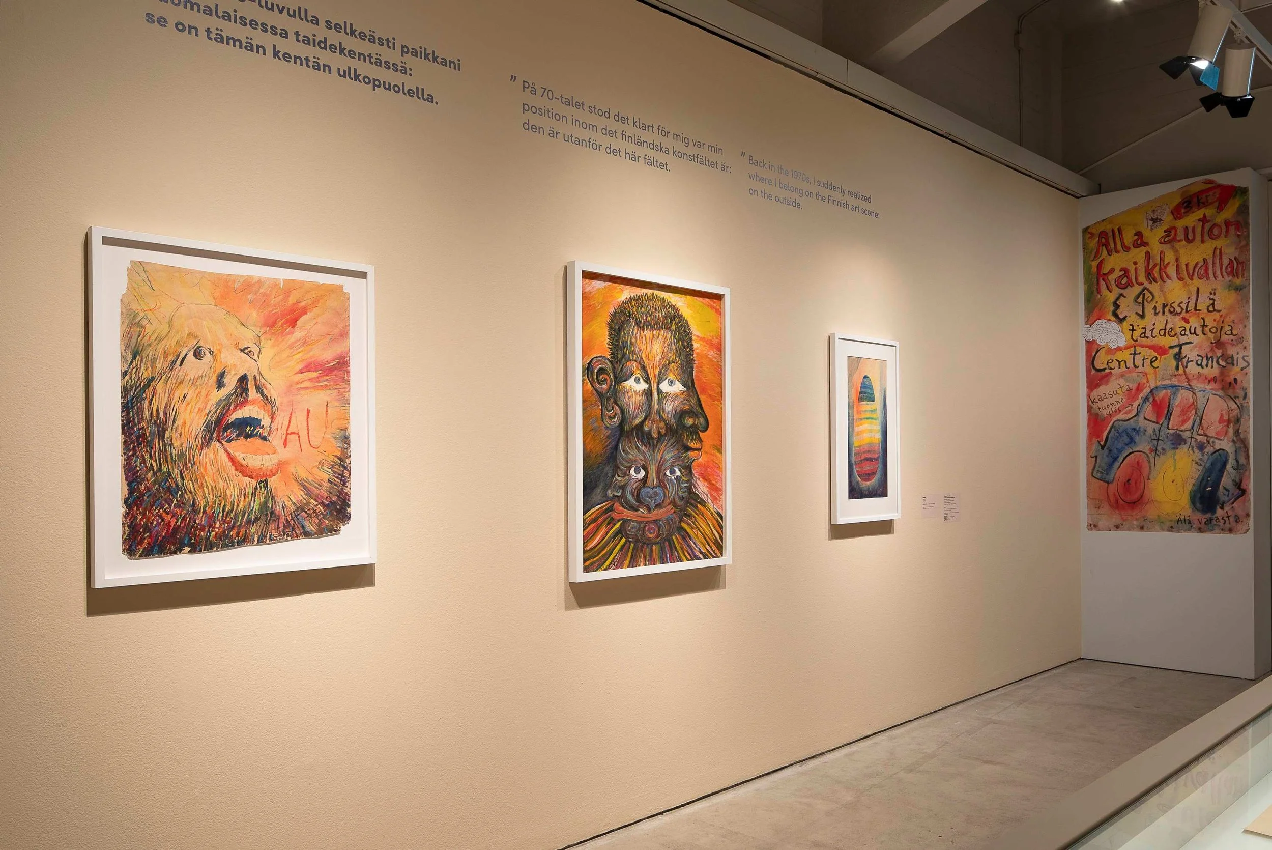 Art gallery wall displaying three vibrant colorful paintings, including a lion with a mane, a face with exaggerated features, and an abstract face, with Finnish text explaining the exhibition.