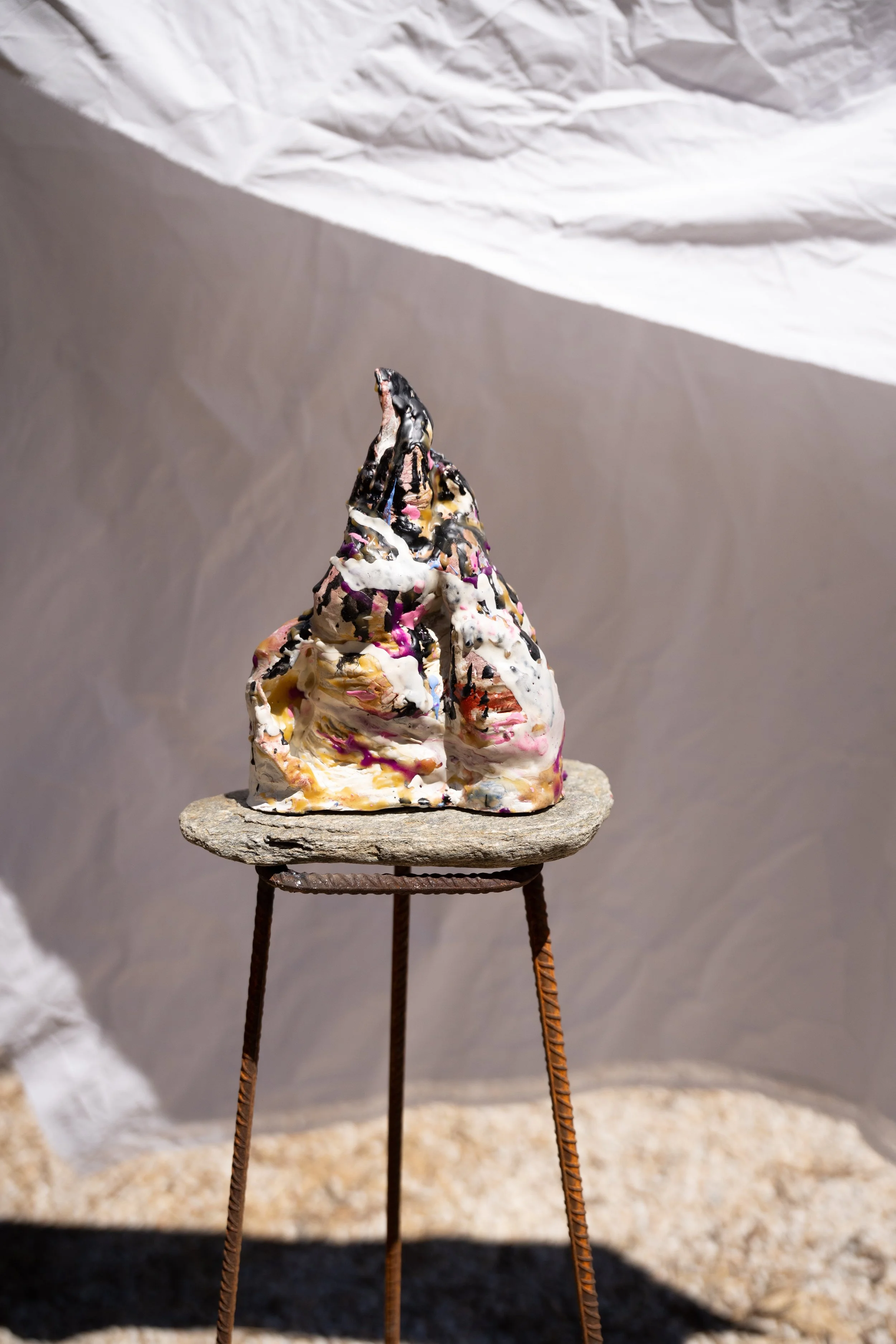 A small sculpture of a mountain, made of painted and textured materials, resting on a round, flat stone, supported by thin, metal rods. The background consists of crumpled and smooth paper. The sculpture resembles a peak with layered colors and textu