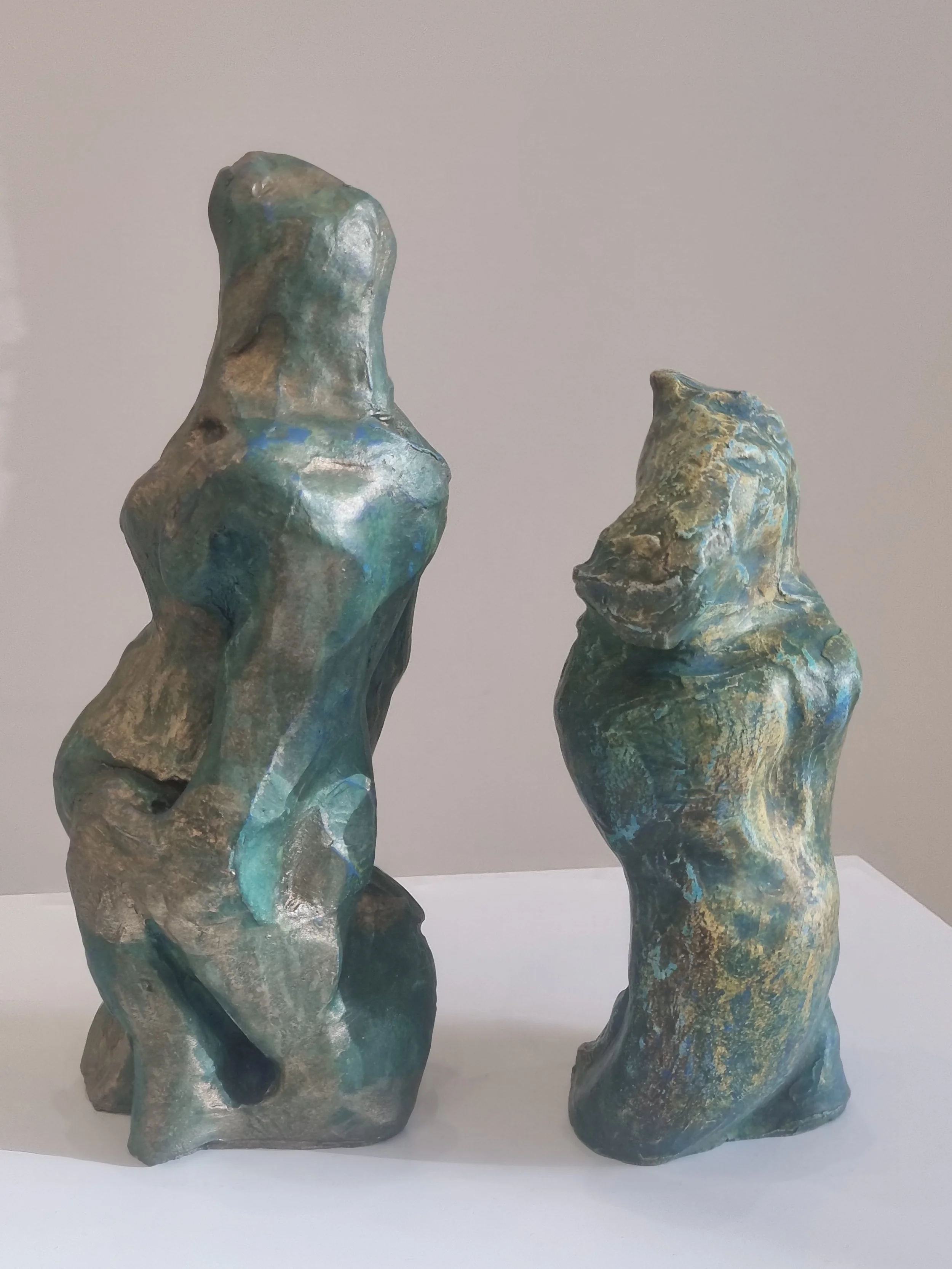 Two abstract sculptures resembling organic shapes or figures, made of metal with a patina finish in shades of green, blue, and bronze, displayed on a white surface against a plain light gray background. Ceramic sulpture by Finnish artist Satu Metsola
