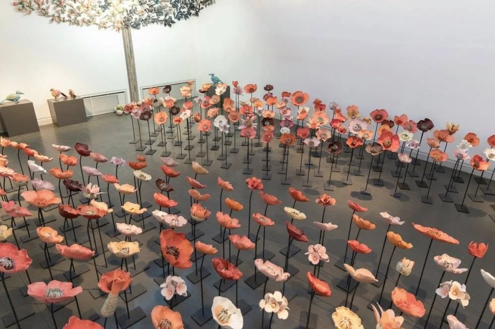 An art exhibit featuring numerous handcrafted ceramic flowers on black metal stands arranged in rows and displayed in a gallery with plain white walls.
