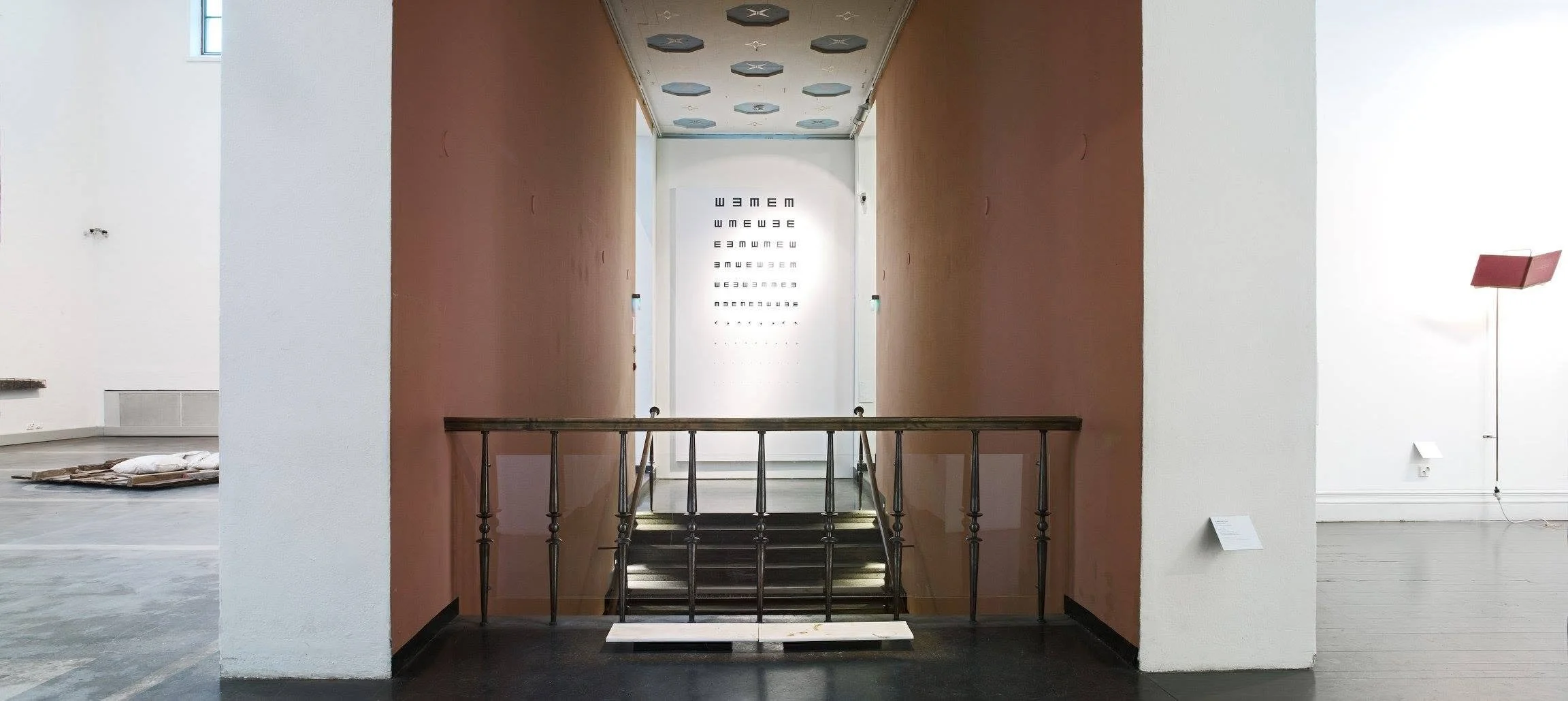 Interior view of an art gallery with a staircase and orange walls. An eye chart is displayed on the wall at the top of the staircase, and a pink floor lamp is on the right side.