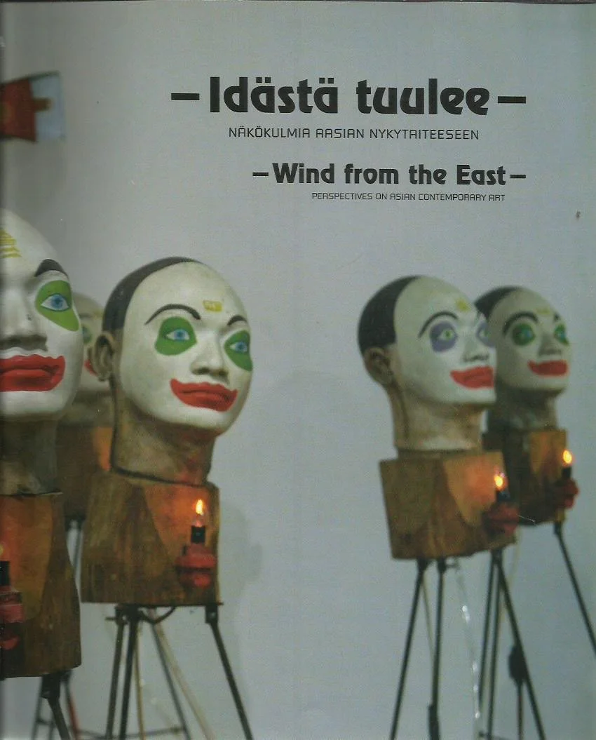 Exhibition display with several large Asian masks on wooden stands, decorated with colorful facial features, against a plain background with informational text about Asian contemporary art and perspectives.