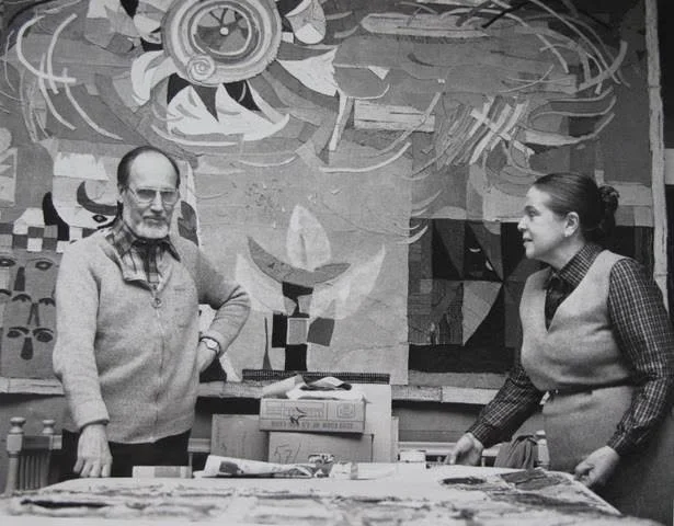 Two women are standing in front of a colorful abstract mural. One woman with glasses and a scarf around her neck is on the left, and the other woman with her hair tied back is on the right. They are looking at each other near a table with art supplies.