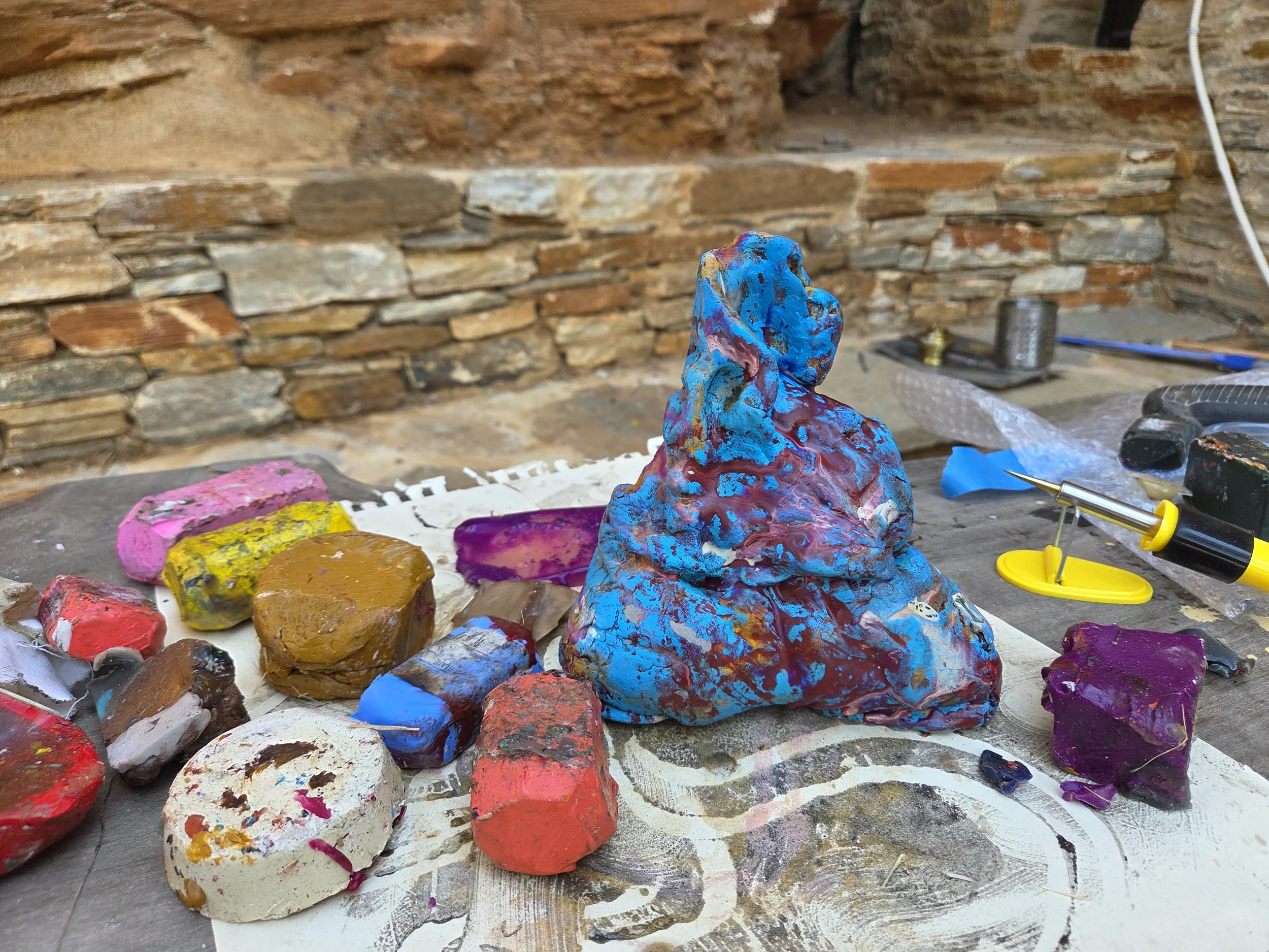 A cluttered worktable with a colorful, textured sculpture in the shape of a seated figure, surrounded by paint splatters, various rocks, and tools, with a brick wall in the background.