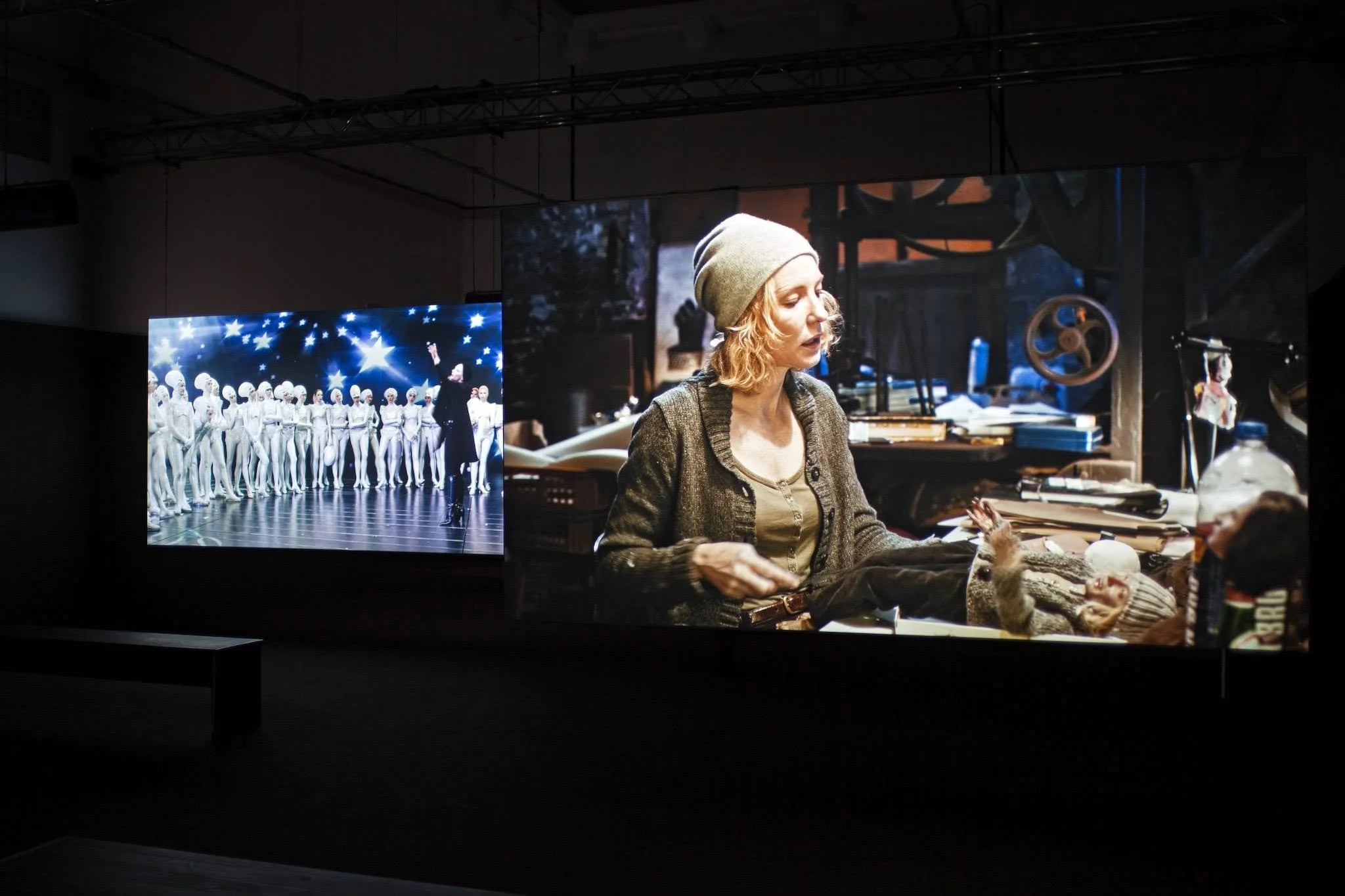A woman with curly blonde hair wearing a beige knit hat and a gray cardigan sitting at a cluttered desk. In the background, two screens display images of ballet dancers; one shows a group of dancers in white costumes on stage, and the other shows a woman in a dark outfit. The surroundings include various objects and equipment related to artistic work.
