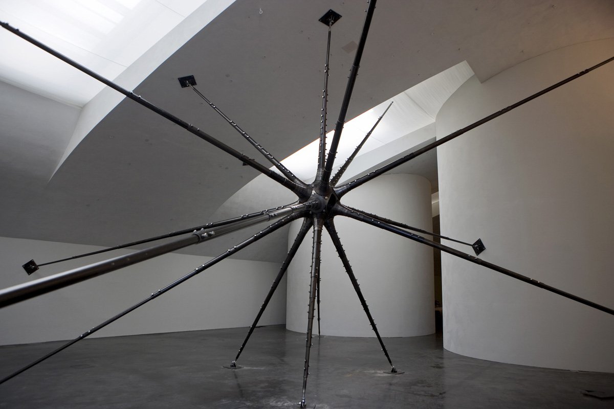 A large, modern sculpture resembling a star with multiple elongated black metal arms radiating from a central sphere, situated in a minimalist gallery space with curved white walls and a polished concrete floor.