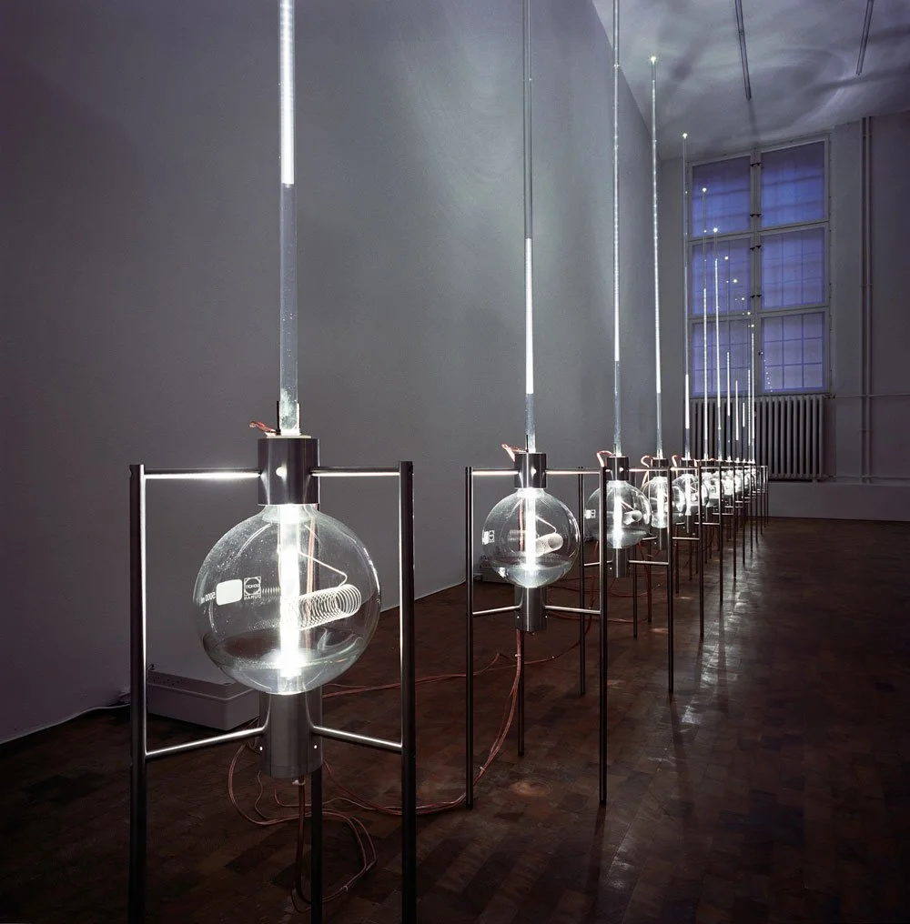 Multiple hanging art installations resembling large test tubes with metallic tops, arranged in a row inside an art gallery with wooden floors and a high window with blue tinted glass.