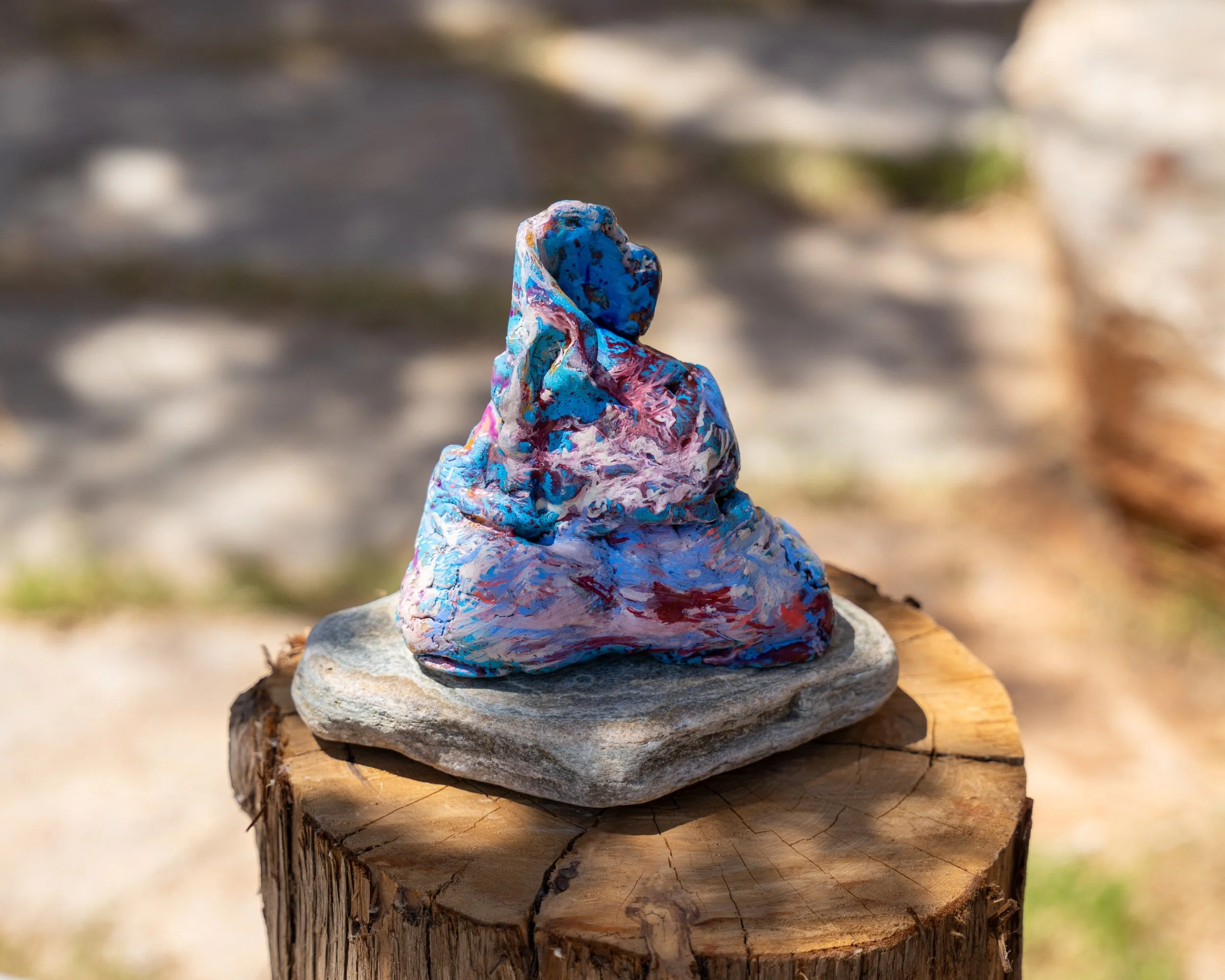 A small colorful painted rock sculpture on a flat rock, placed on top of a tree stump. Abstract sulpture by Finnish artist Satu Metsola