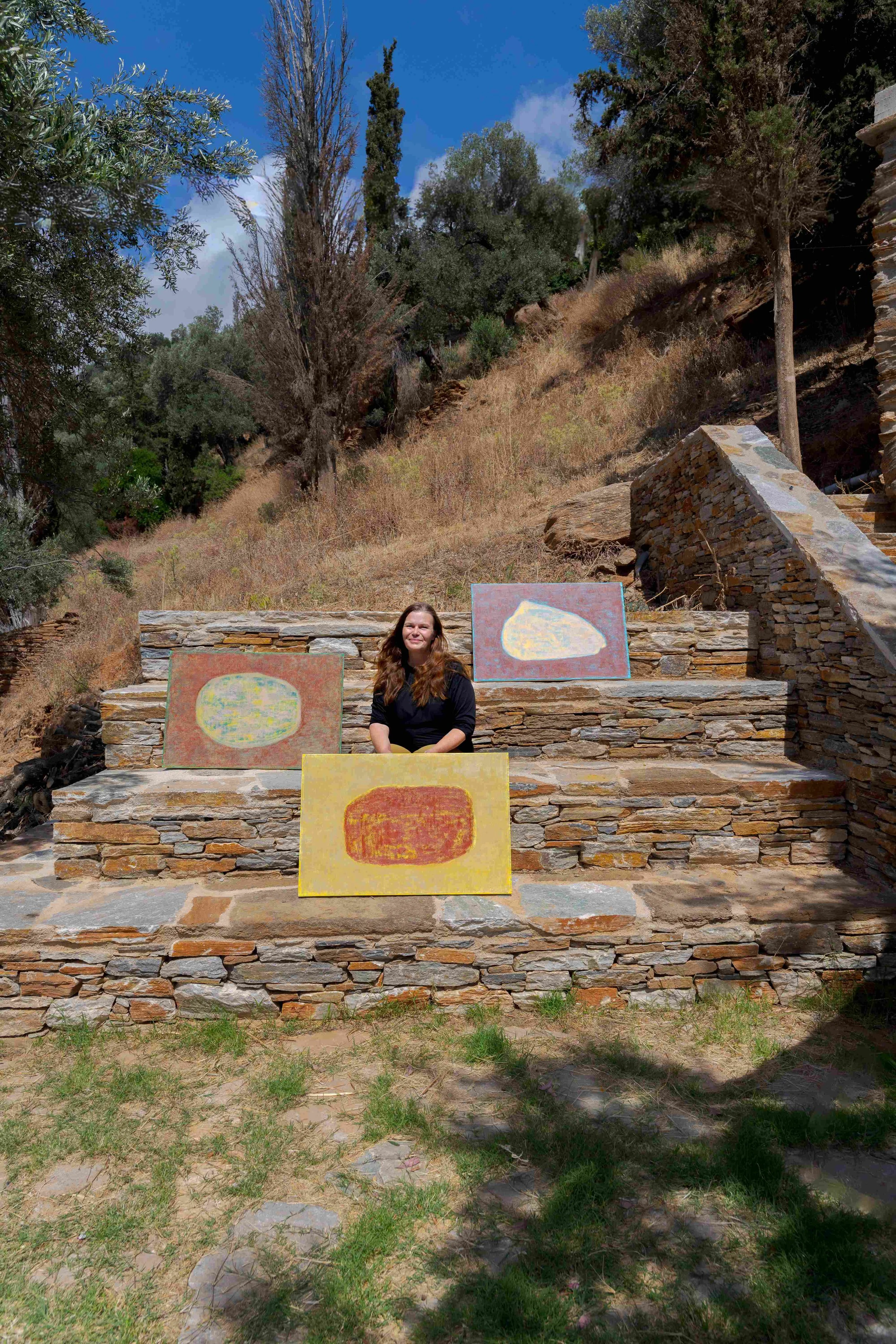 A woman sitting on stone steps outdoors, surrounded by trees and shrubs, with paintings of abstract shapes on the steps. Finnish artist Satu Metsola in Greece with her paintings.