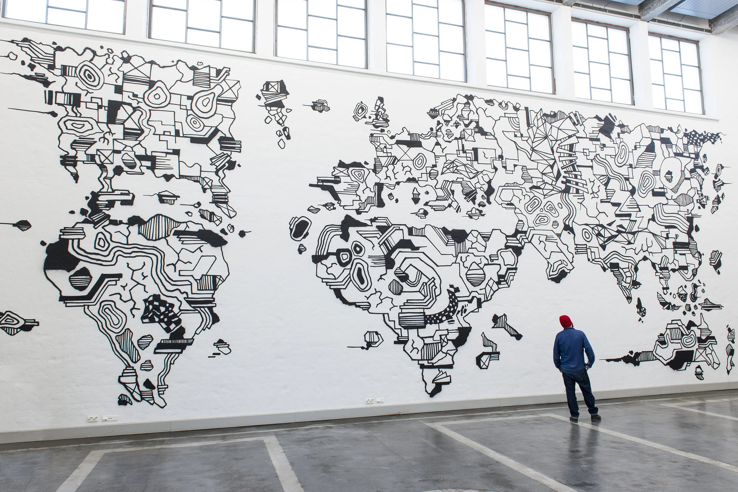 A large, abstract black and white world map mural on a white wall in an industrial-style space with large windows. A person wearing a blue jacket, red hat, and jeans is standing in front of the mural, looking at it.