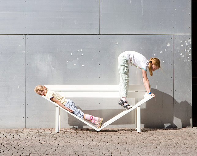 Two children playing on a bench that is tilted and designed for swinging, against a gray concrete wall. The girl on the left is lying on the bench, leaning back, while the girl on the right is balancing on top of the bench, reaching down.