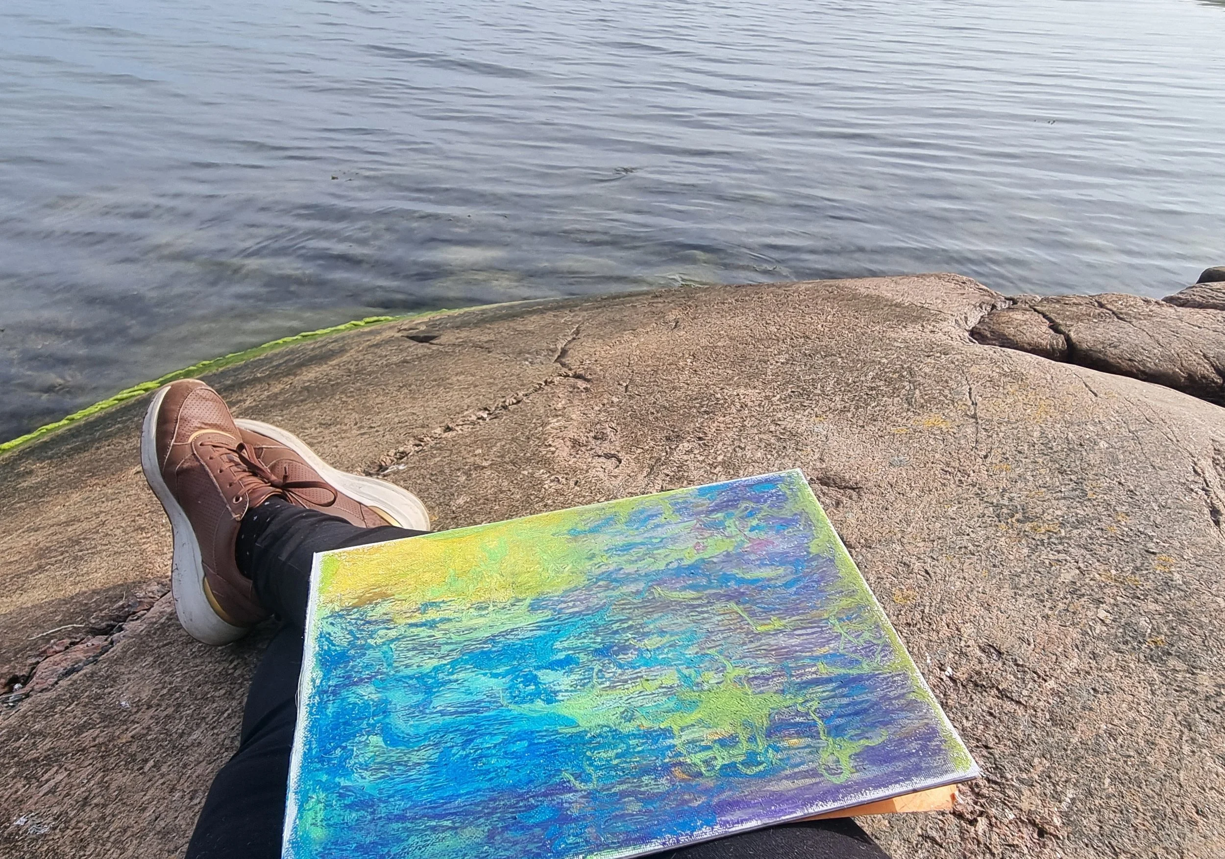 A person sitting on a rocky shoreline with a colorful painted canvas, wearing sneakers and dark pants, with a body of water in the background.