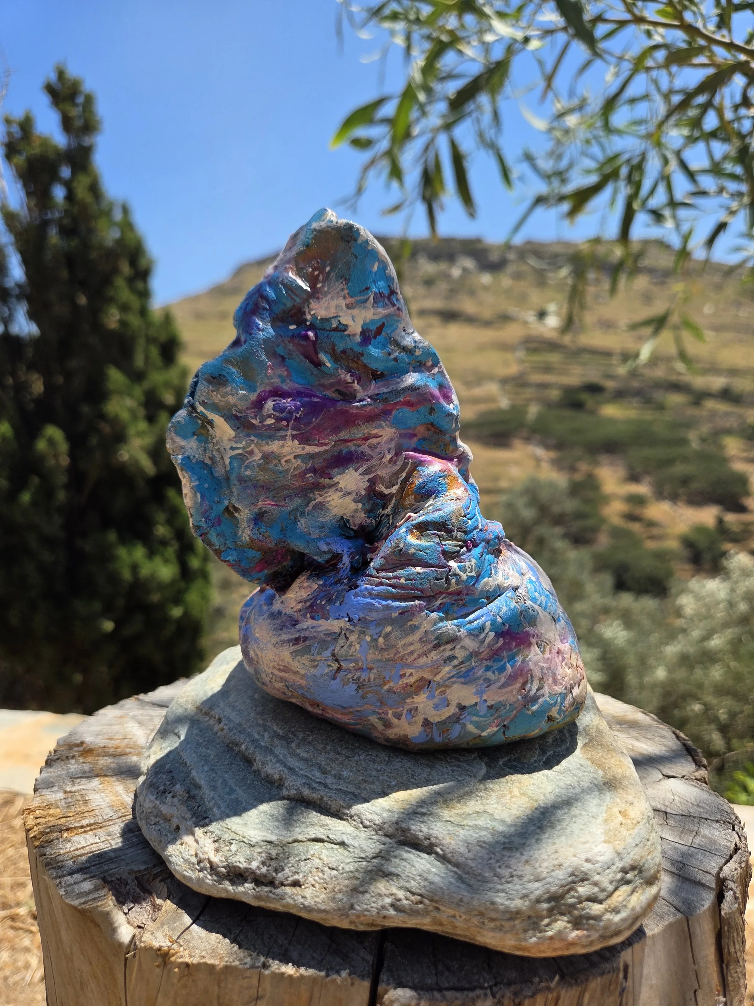 Colorful art work resting on a weathered tree stump outdoors with a mountain, trees, and blue sky in the background. Abstract ceramic sulpture by Finnish artist Satu Metsola
