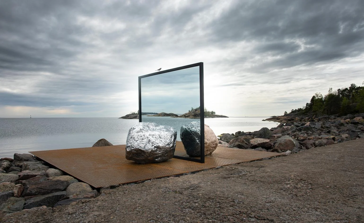 An outdoor art installation by the water featuring large rocks on a metal platform, with a metal frame mirror reflecting rocks and a cloudy sky, overlooking a rocky shoreline and a distant small island.