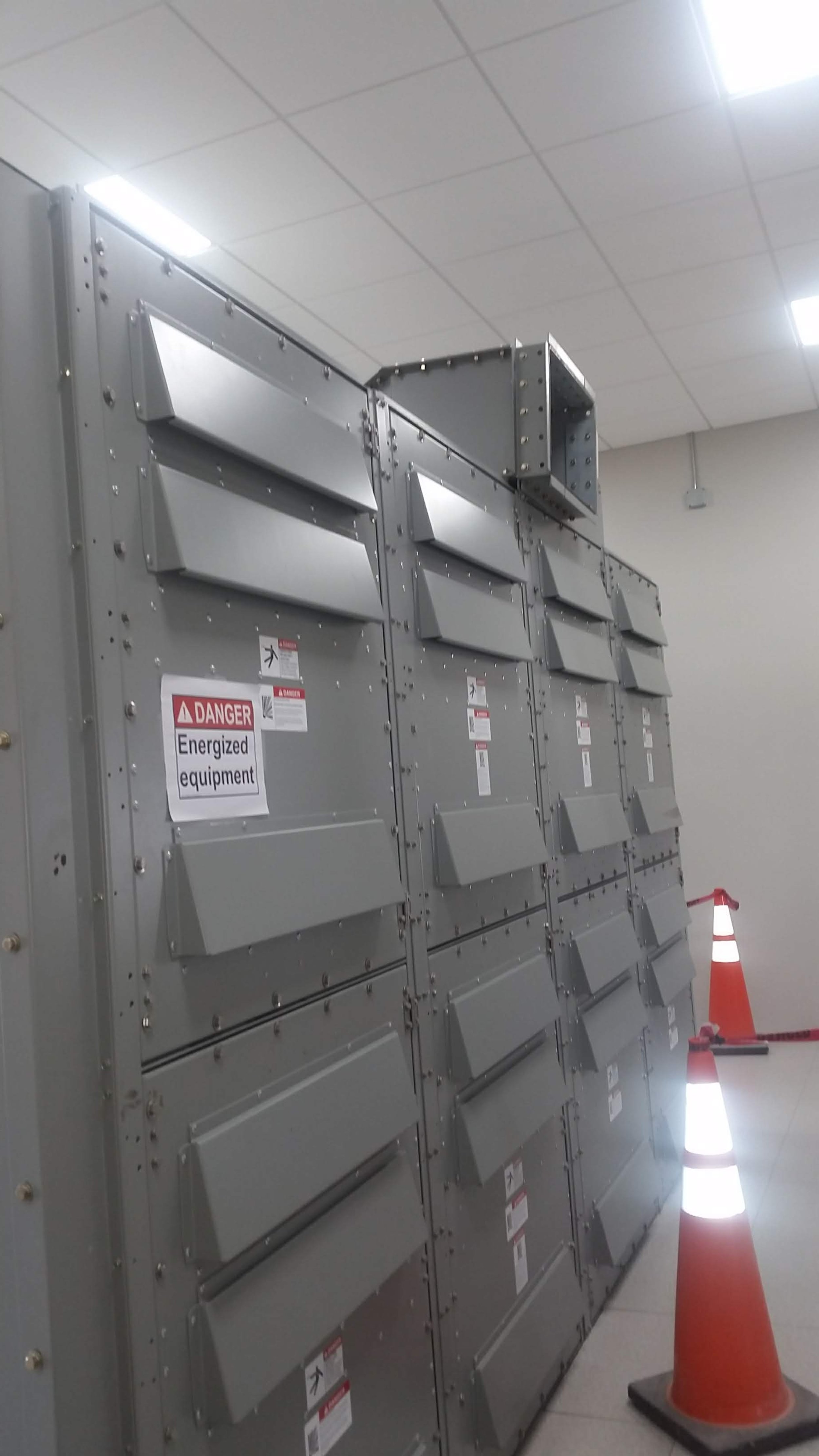 Large gray electrical equipment with warning labels and orange traffic cones nearby in an indoor setting.