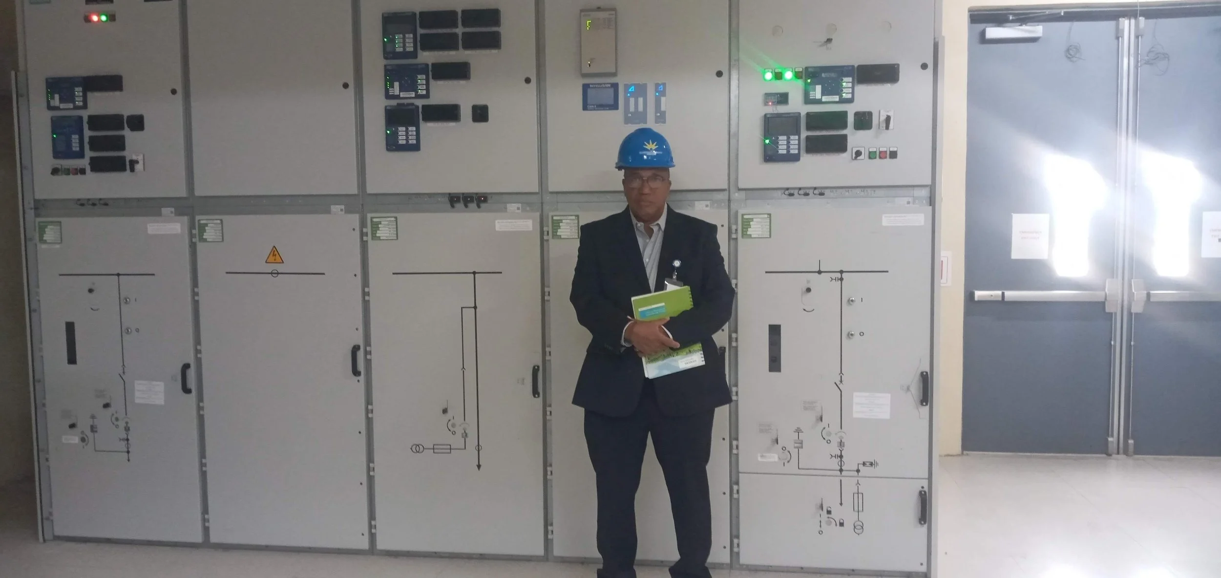 Leecorp Ltd's Founder and CEO, Francois Lee, in a business suit and a blue safety helmet stands in front of electrical control panels in an industrial facility.