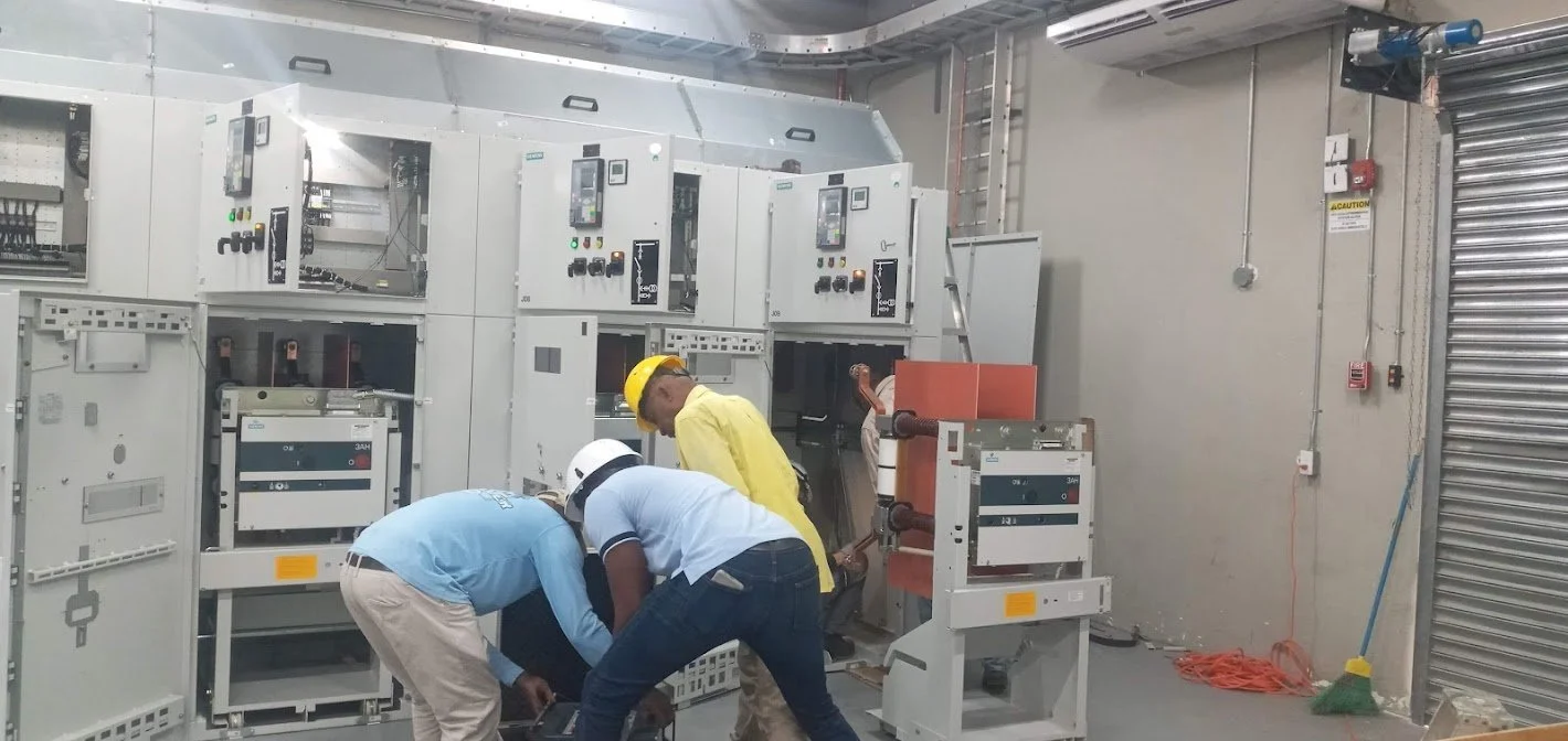 Three workers wearing helmets and safety equipment working inside an industrial electrical or control panel room. They are bent over, examining or working on electrical components.