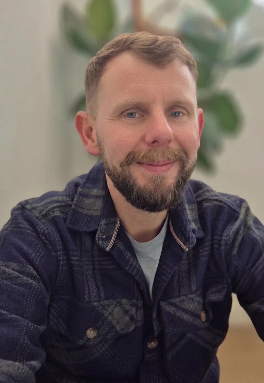 A man with short light brown hair, a beard, and a smirk, sitting indoors in front of a green plant. He is wearing a dark plaid shirt over a gray t-shirt.