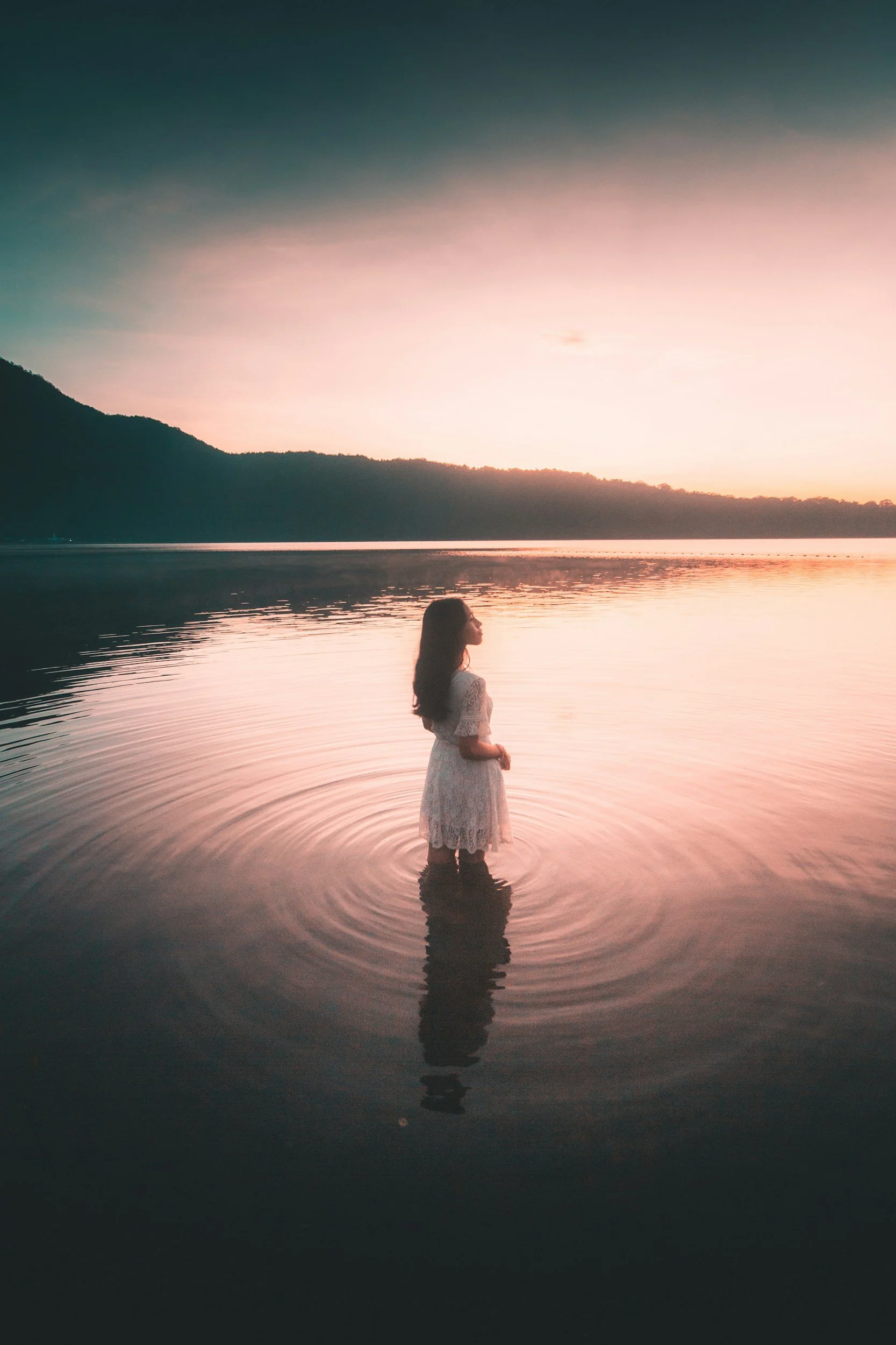 A woman standing in shallow water at sunset or sunrise with mountains in the background and a pink, orange, and blue sky.