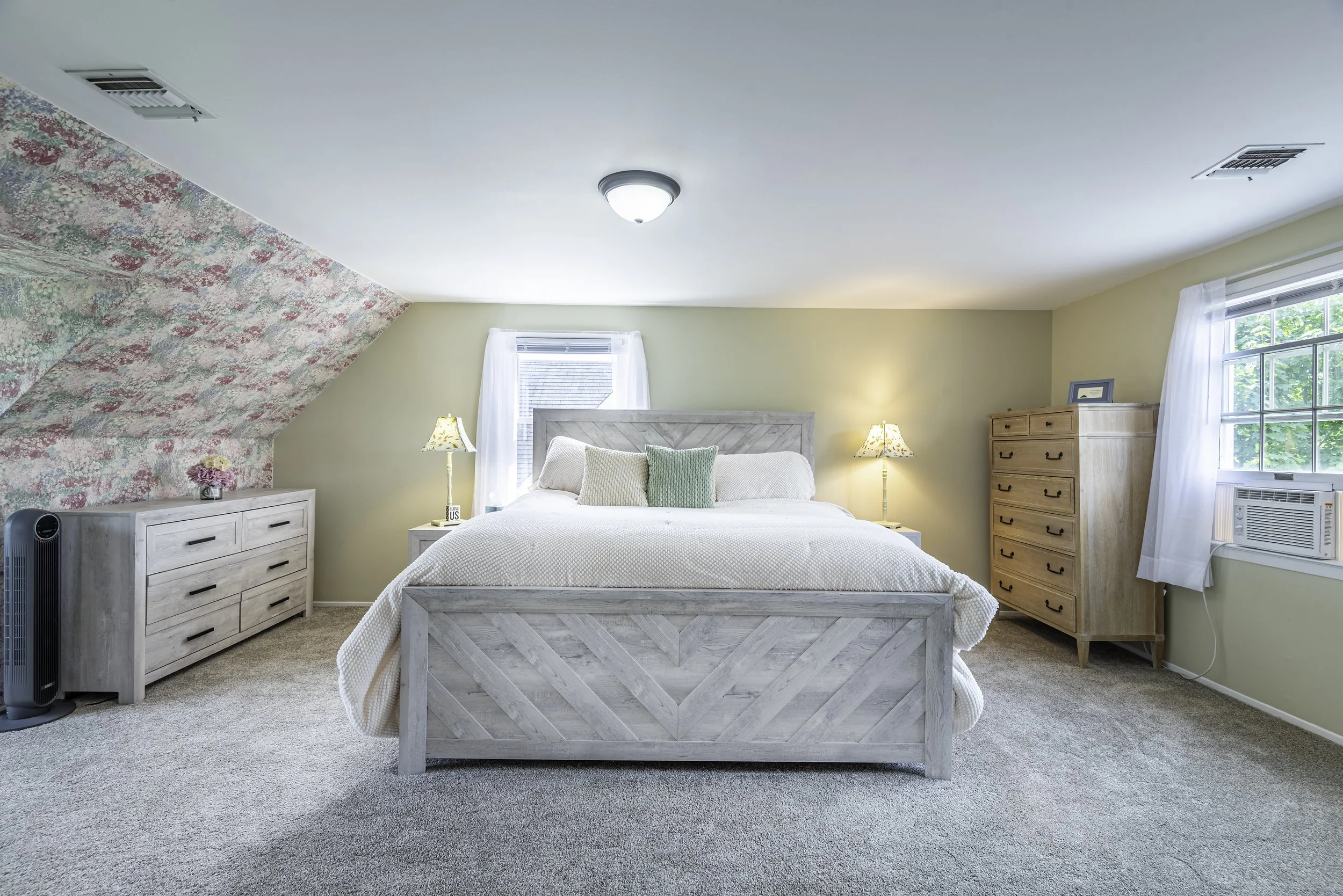 Spacious primary bedroom with cozy carpet flooring, natural light, stylish wood furniture, floral accent wall, and a large bed centered in the room.