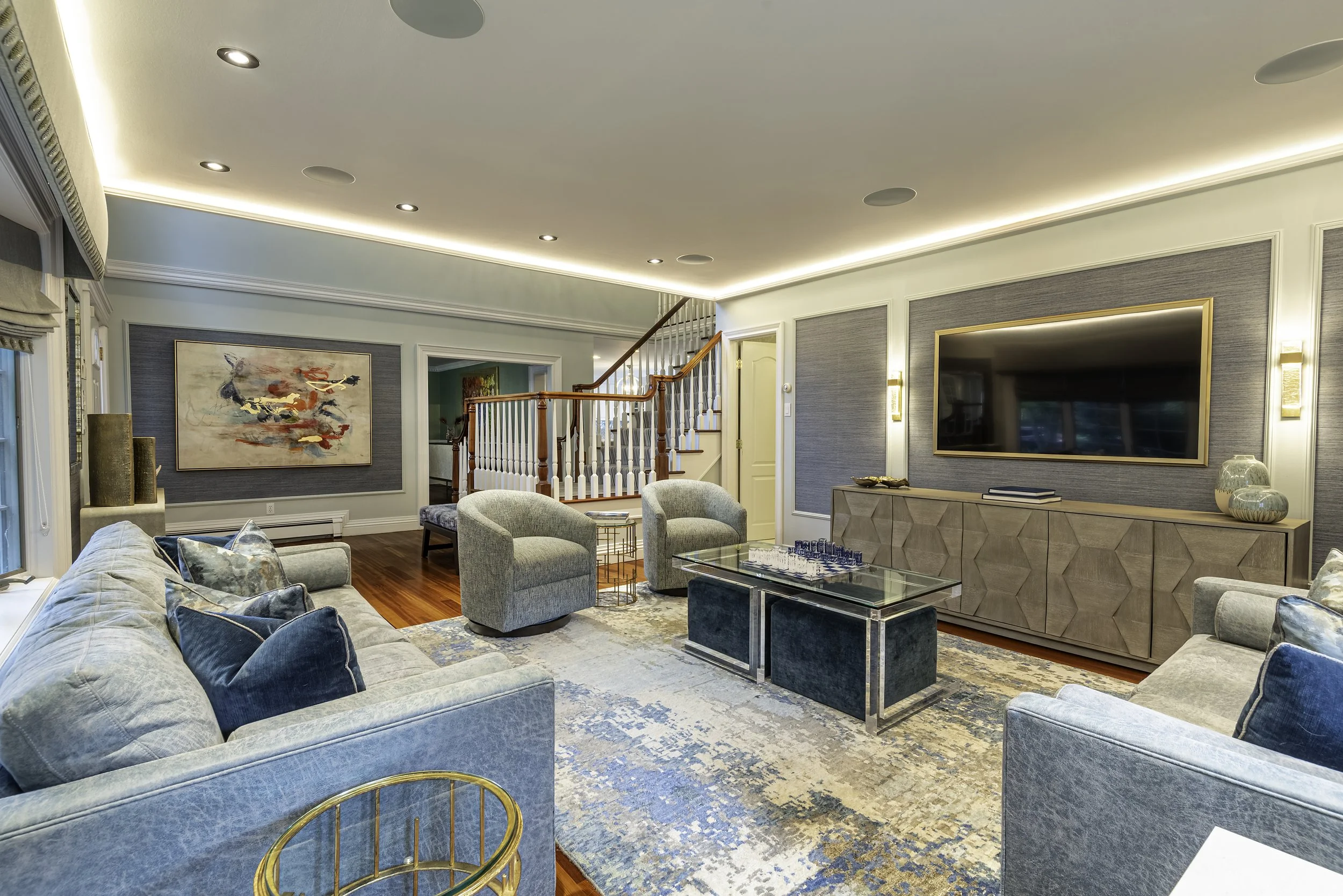 Living room with a large television mounted on the wall, a beige sideboard, a glass coffee table, and multiple sofas and armchairs with blue and gray cushions. There is an abstract painting on the wall, hardwood floors, and a staircase with a wood railing in the background.