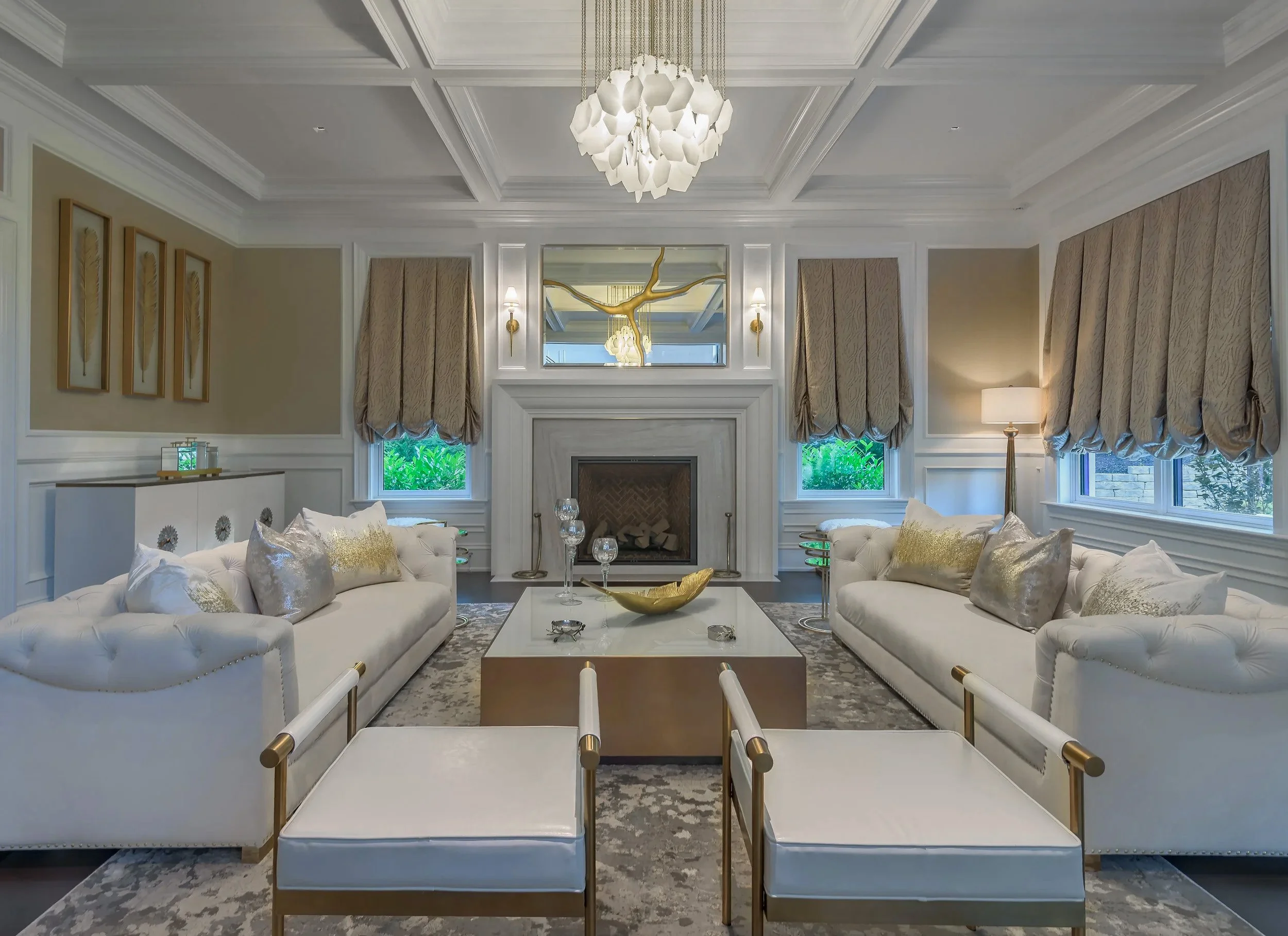 Elegant living room with white sofas, gold and silver pillows, a central coffee table with decorative items, a fireplace, windows with beige curtains, and a modern chandelier.