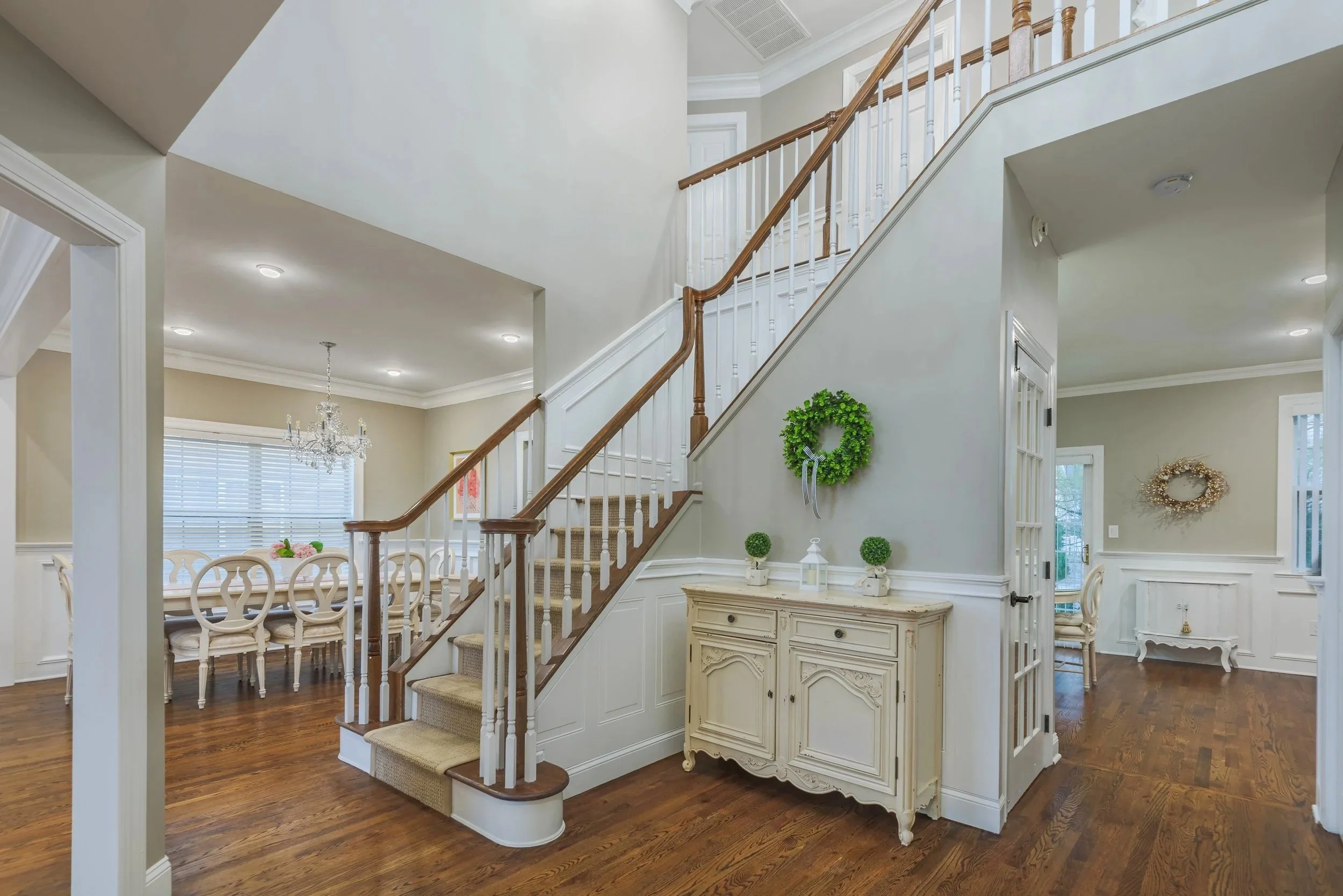 Interior view of a home with a staircase, dining area with a chandelier, and wall decorations with wreaths. Hardwood floors, cream-colored walls, and white trim.