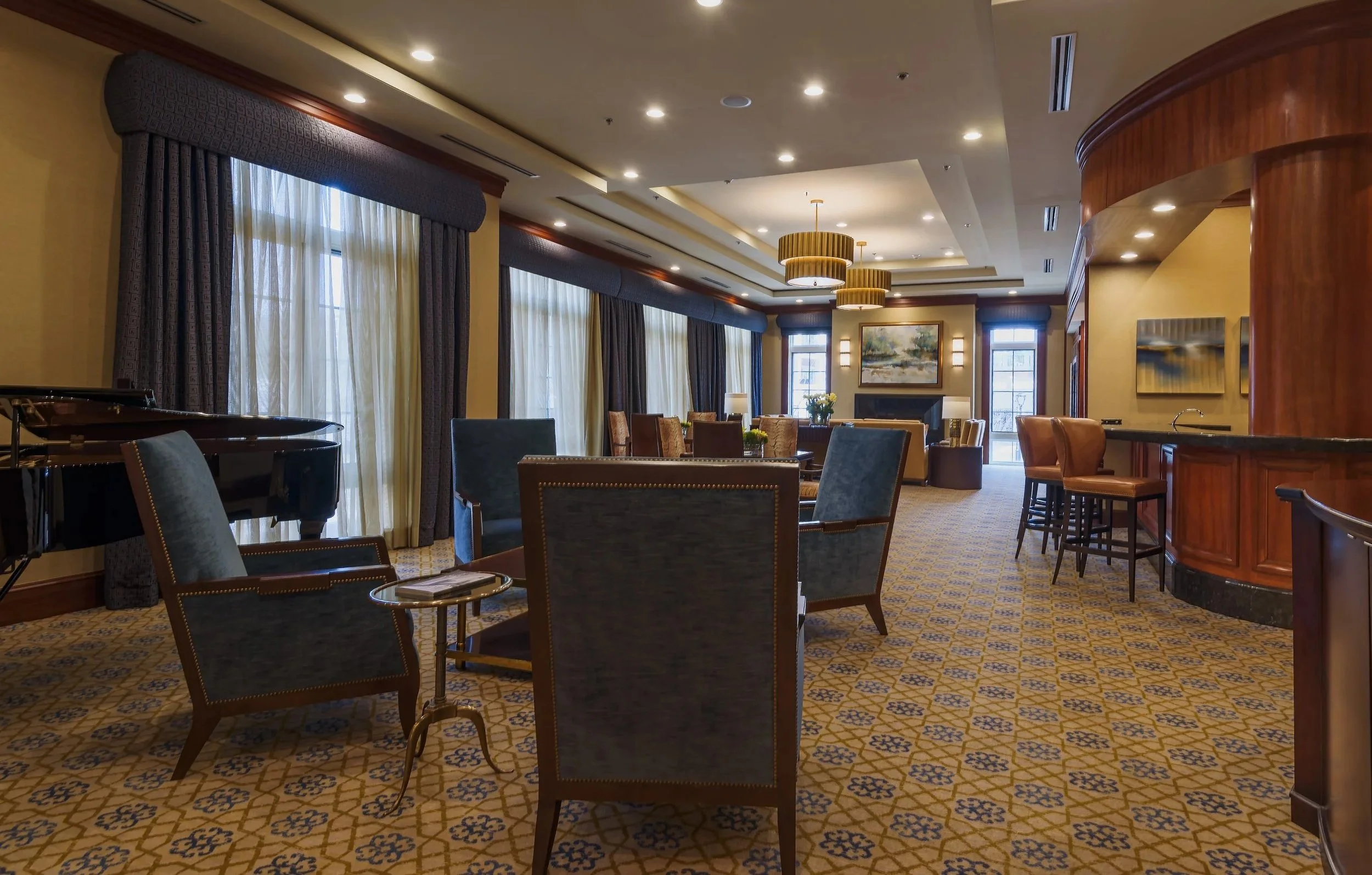 Elegant hotel lounge with large windows, velvet curtains, plush armchairs, a grand piano, and modern artwork, illuminated by ceiling lights and hanging chandeliers.
