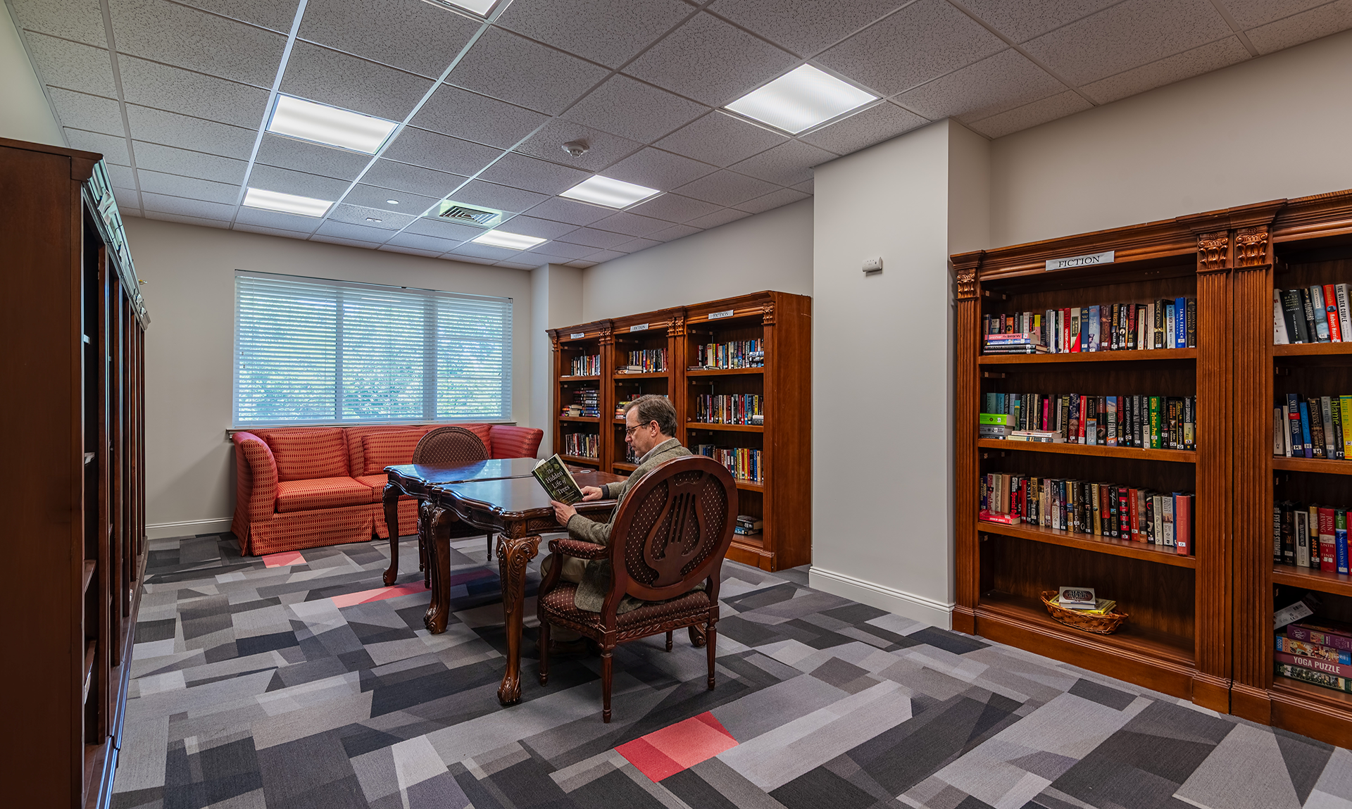 A person sitting and reading in a well-lit library or study room with wooden bookshelves filled with books, a red couch by a window, and a patterned carpet.