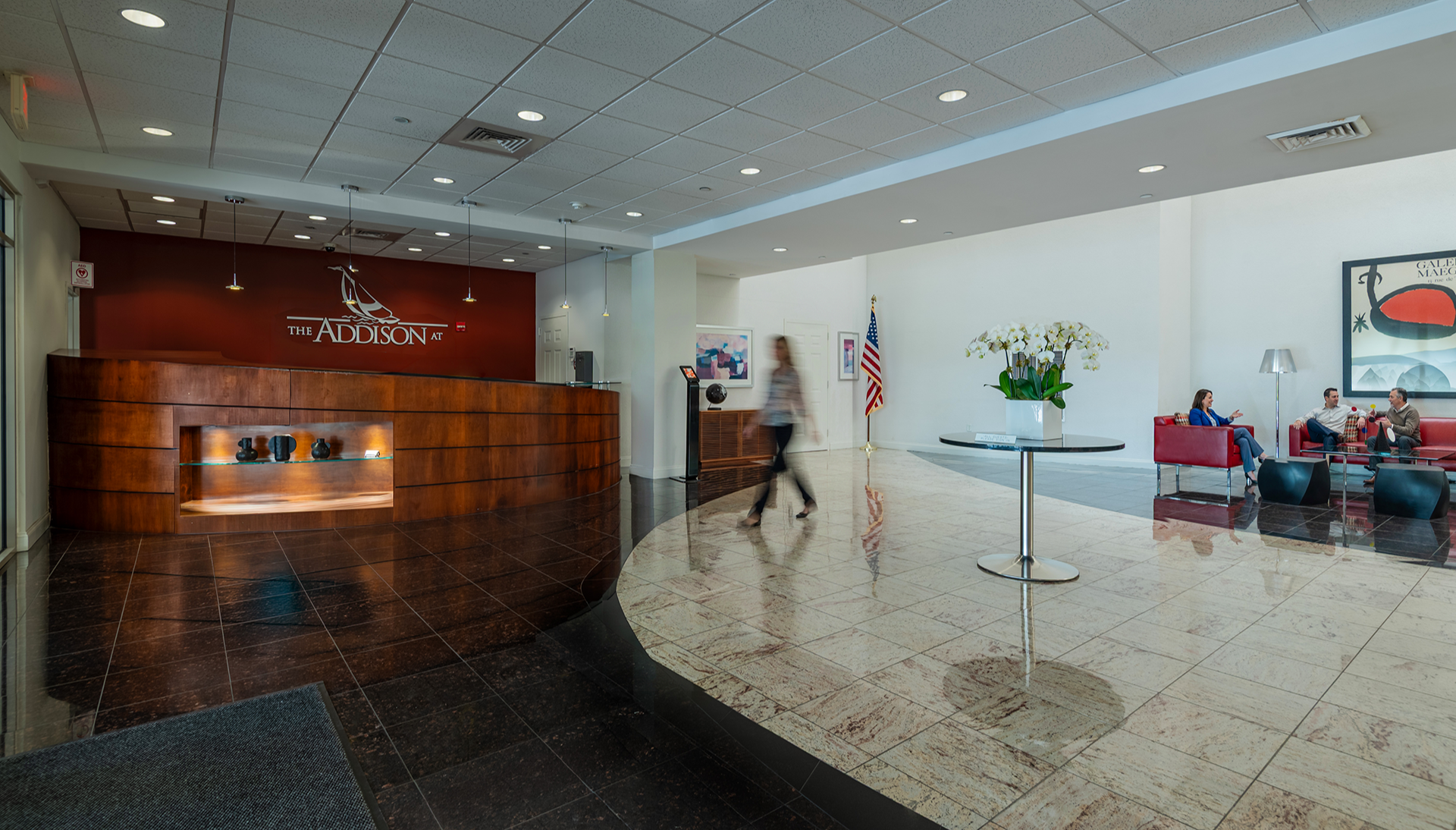 Hotel lobby with a curved wooden reception desk on the left and a seating area on the right screened by a glass partition. The seating area has red couches, a black coffee table, and a tall floor lamp, with people sitting and talking. A large flowerp