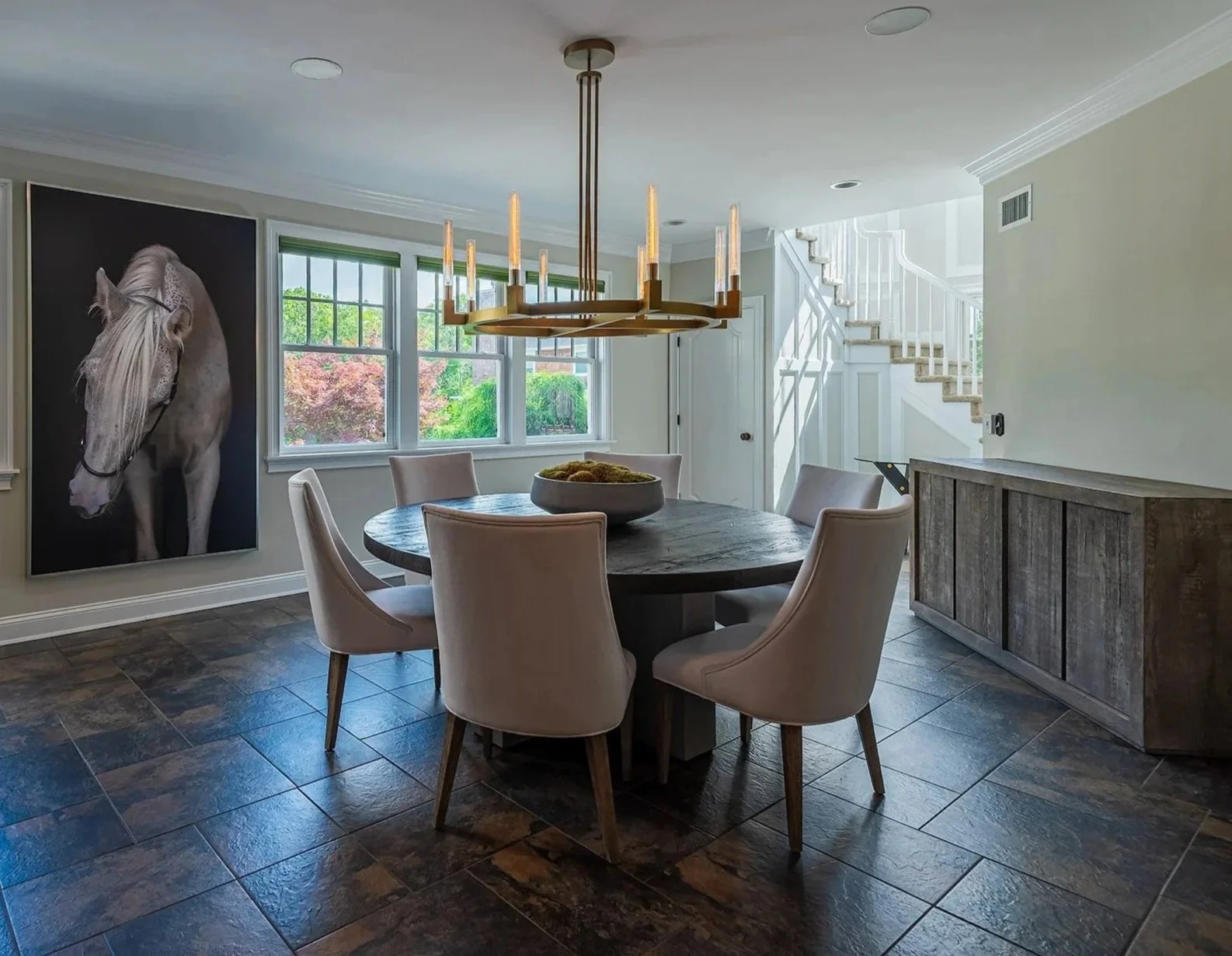 Dining room with round wooden table surrounded by six beige upholstered chairs. On the wall is a large portrait of a white horse. Natural light from multiple windows reveals a view of lush green trees outside. A modern chandelier hangs above the tabl