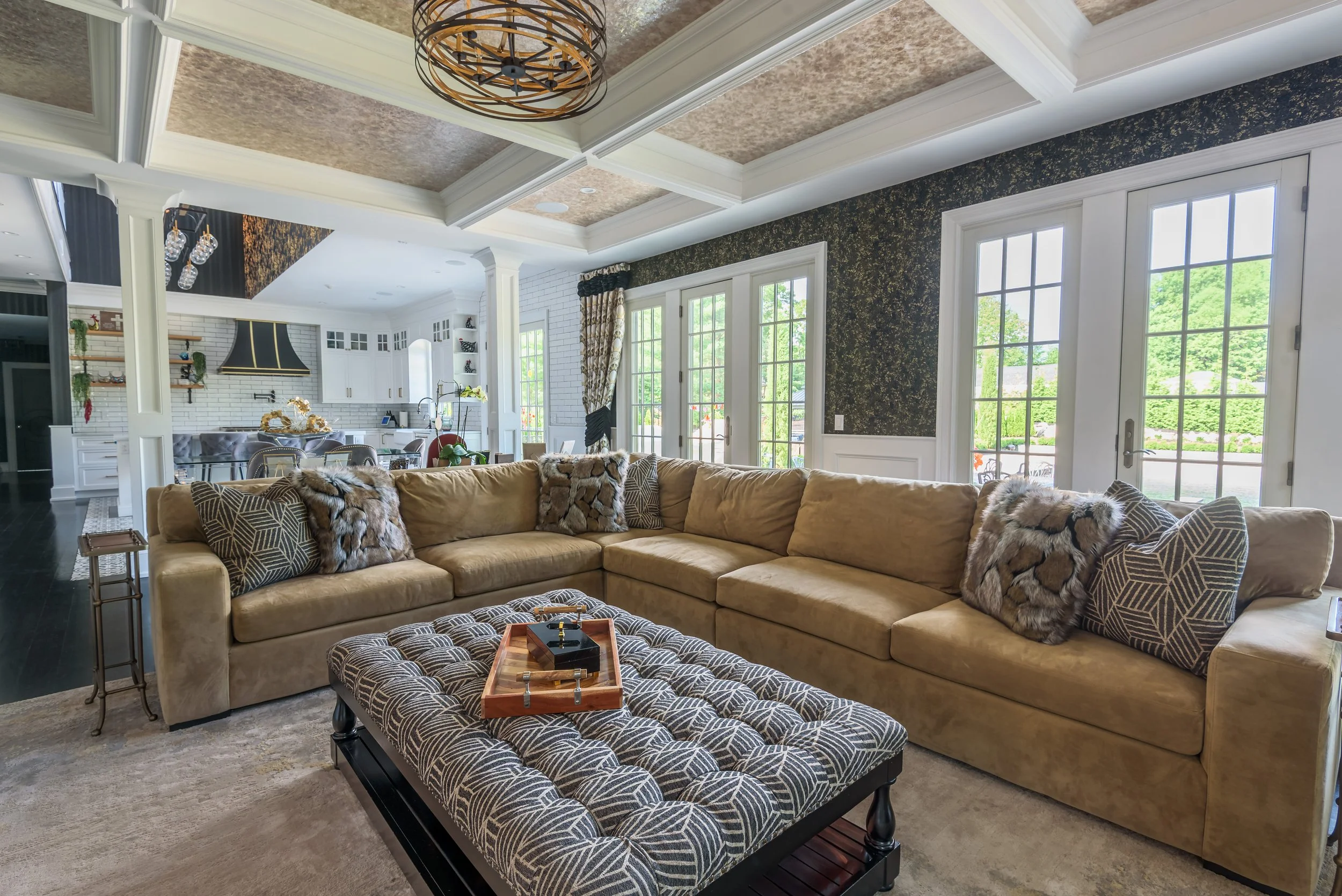 Architectural interior photography of an open plan living room and kitchen featuring coffered ceiling, sectional seating, large windows, and bright natural light.