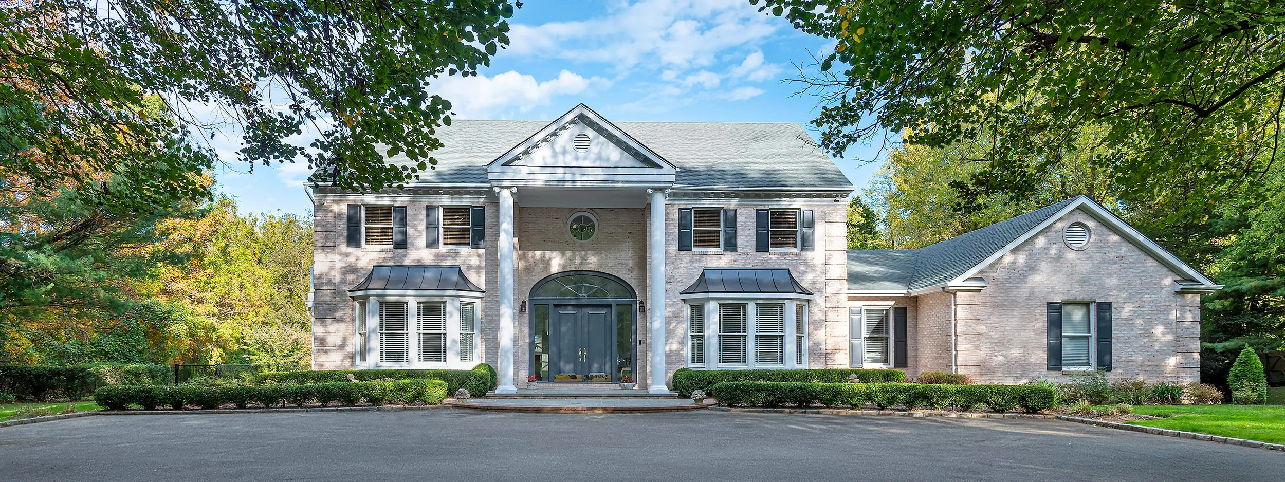   Front exterior of a custom brick colonial home featuring grand columns, arched transom entry, standing seam bay window
  awnings, and professional landscaping — photographed by Palma Design. Architectural photography for luxury builders and
  desig