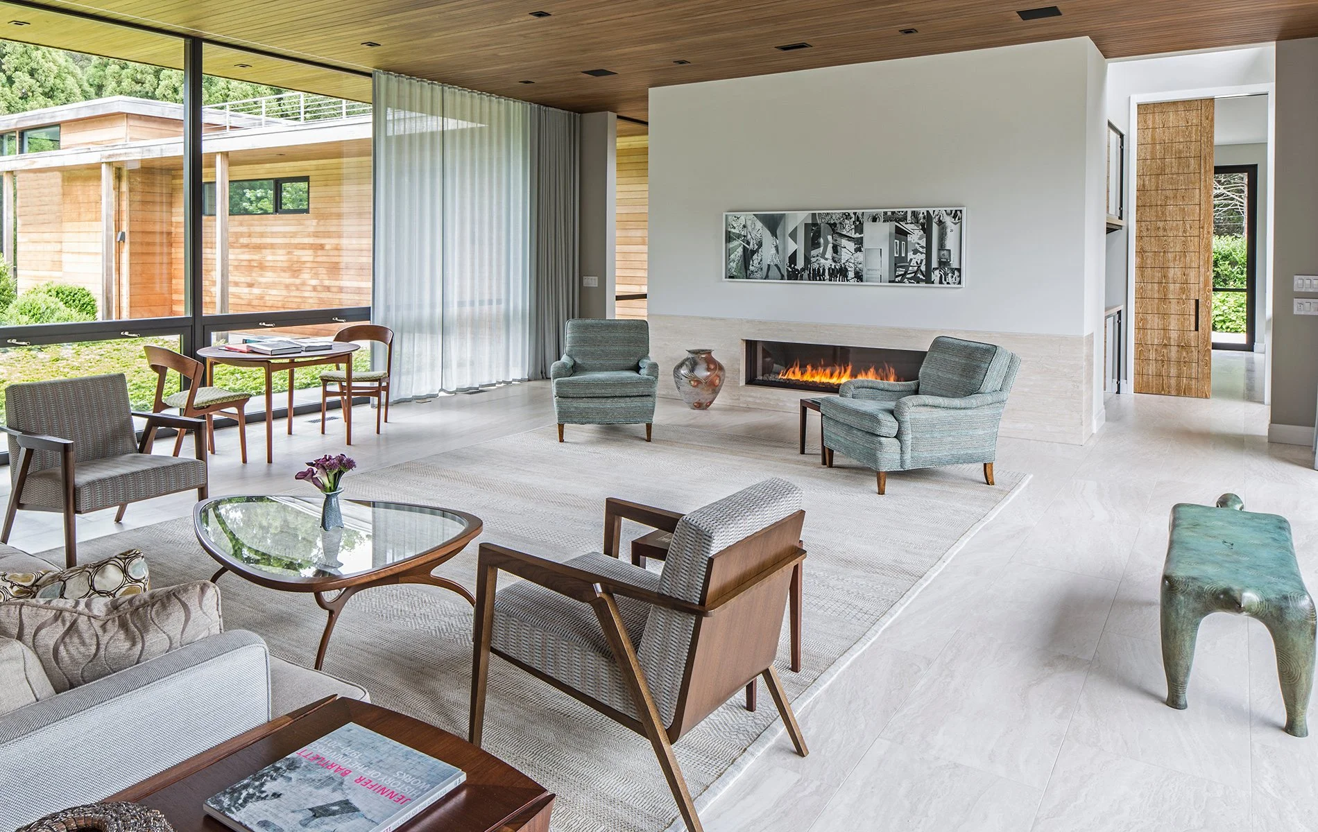 Modern living room with large glass windows, white walls, green armchairs, a fireplace, coffee table, and decorative items.