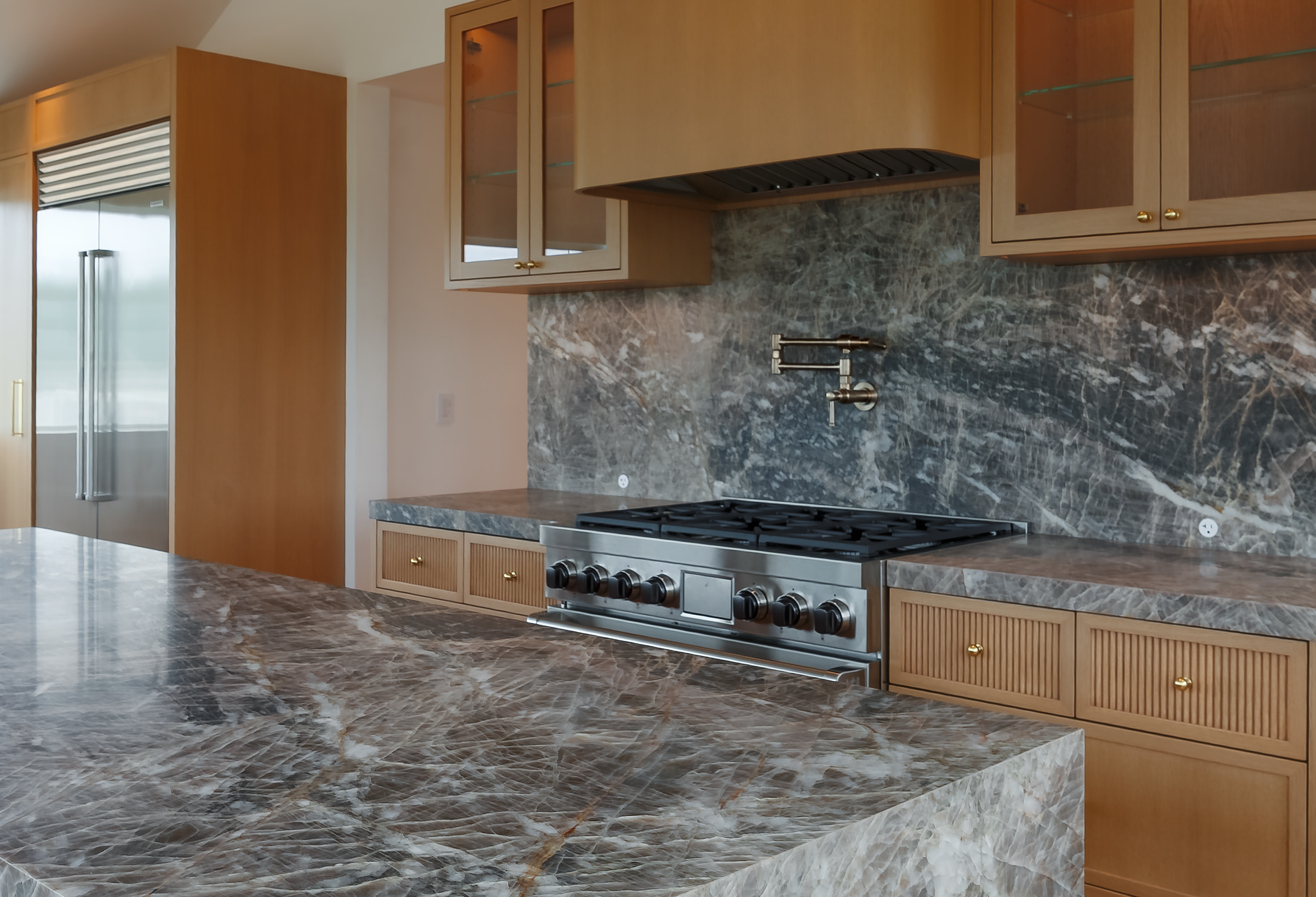 Kitchen detail featuring a stainless steel range, veined stone backsplash and island, brass pot filler, and fluted wood cabinetry.