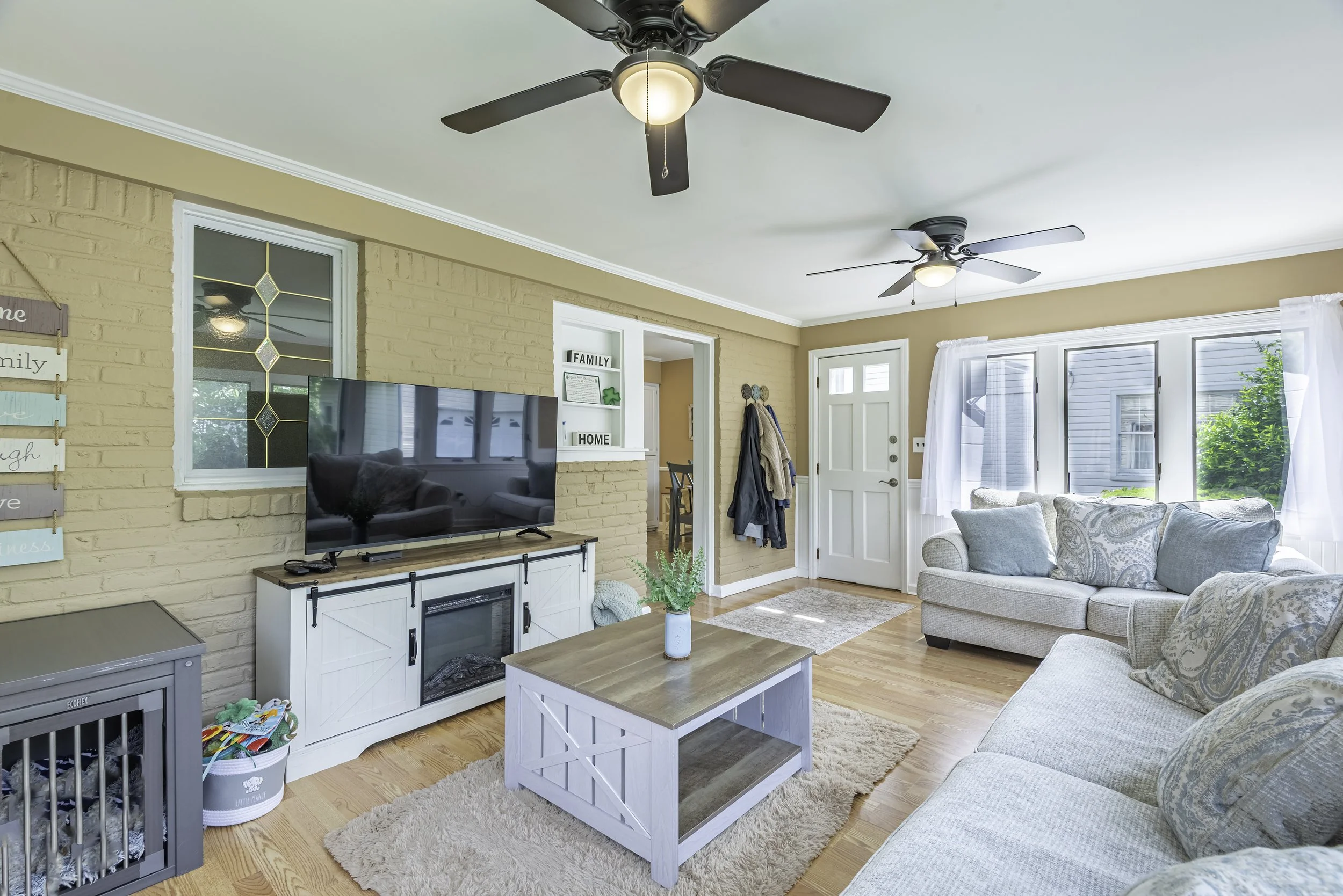 Beautiful interior real estate photo of a family room with hardwood floors, bright windows, ceiling fans, cozy seating, and TV feature wall.