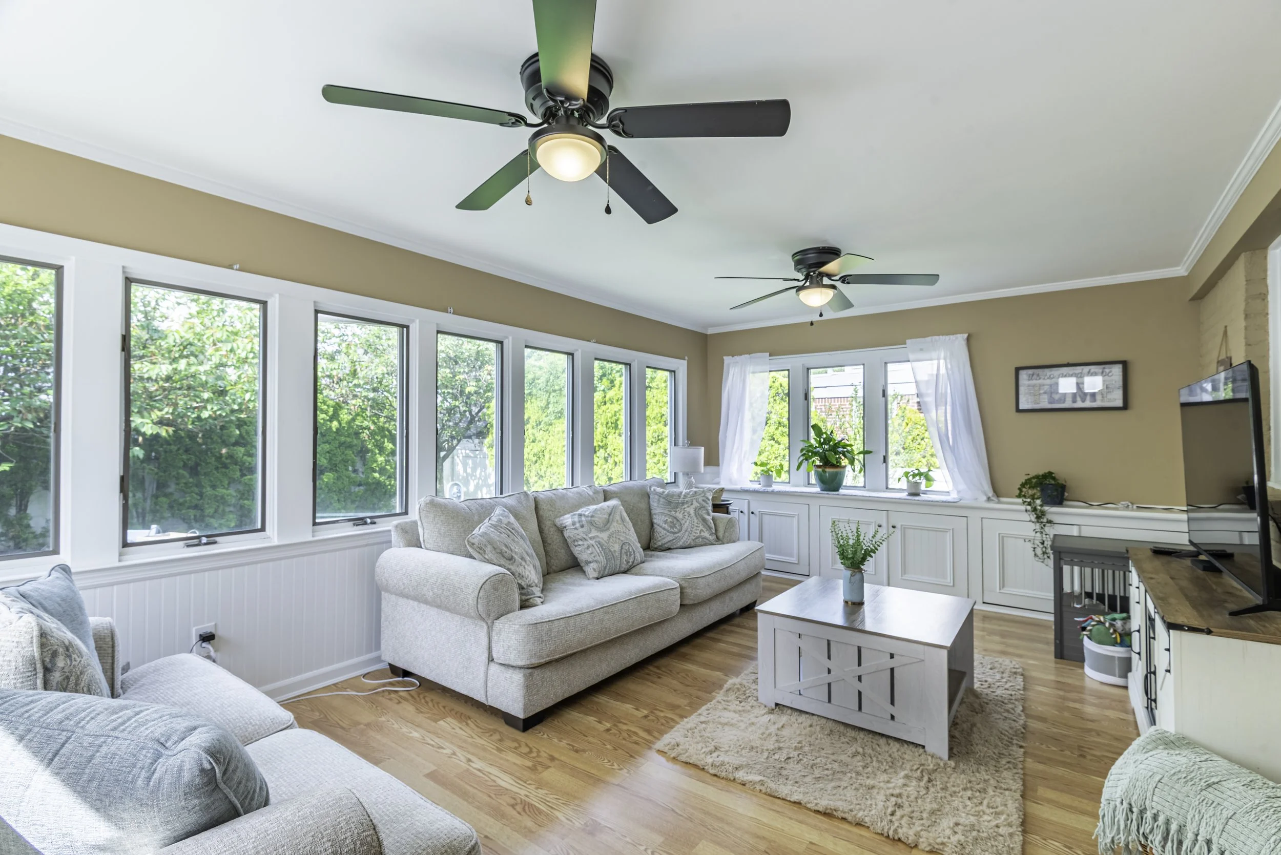 Bright sunroom-style family room featuring large windows, hardwood floors, ceiling fans, cozy seating, and abundant natural light.