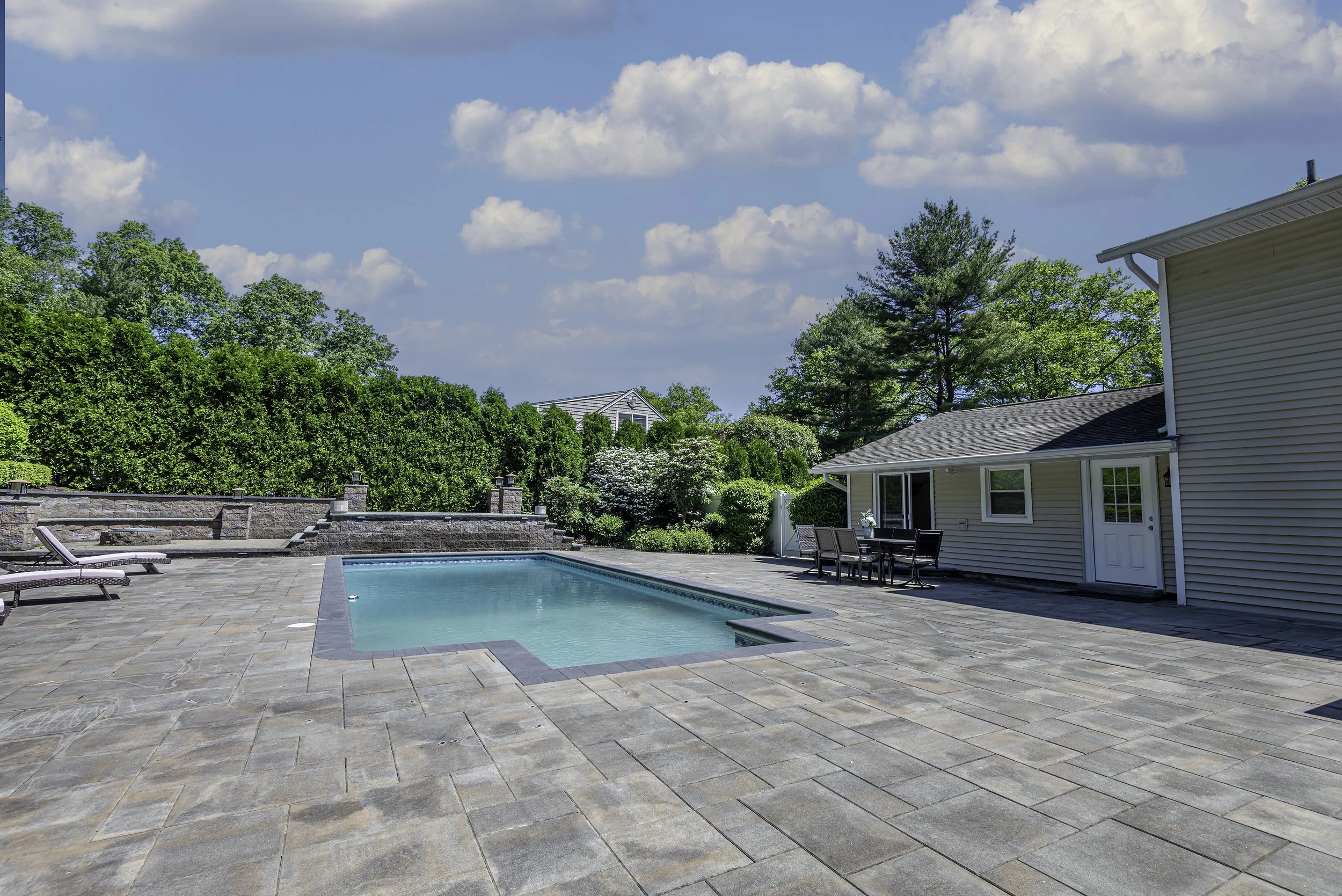 Exterior real estate photo showcasing a spacious backyard with patio, pool, seating area, and landscaped greenery.