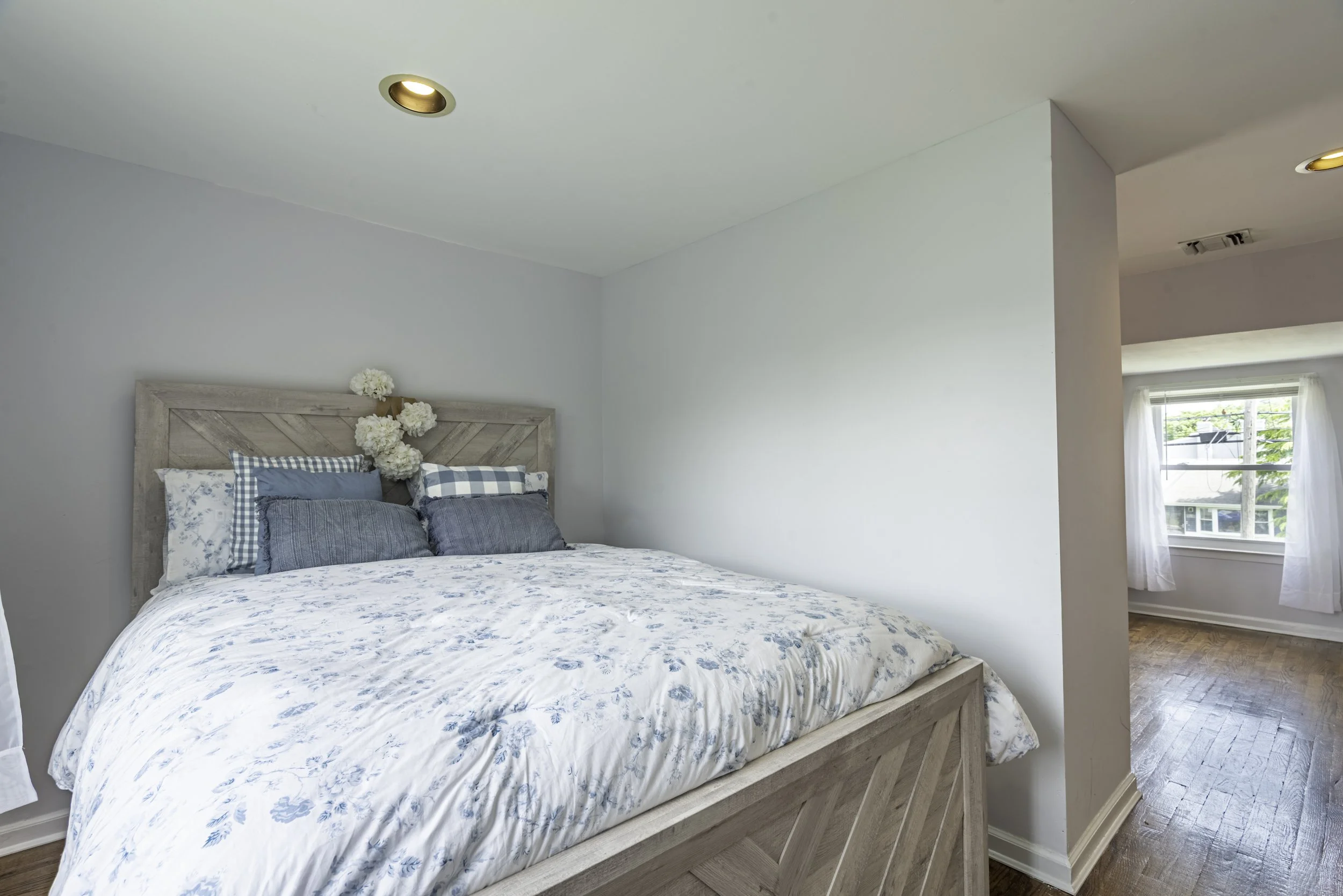 Primary bedroom with wood bed frame, soft neutral walls, cozy bedding, recessed lighting, and adjoining sitting area with natural light.