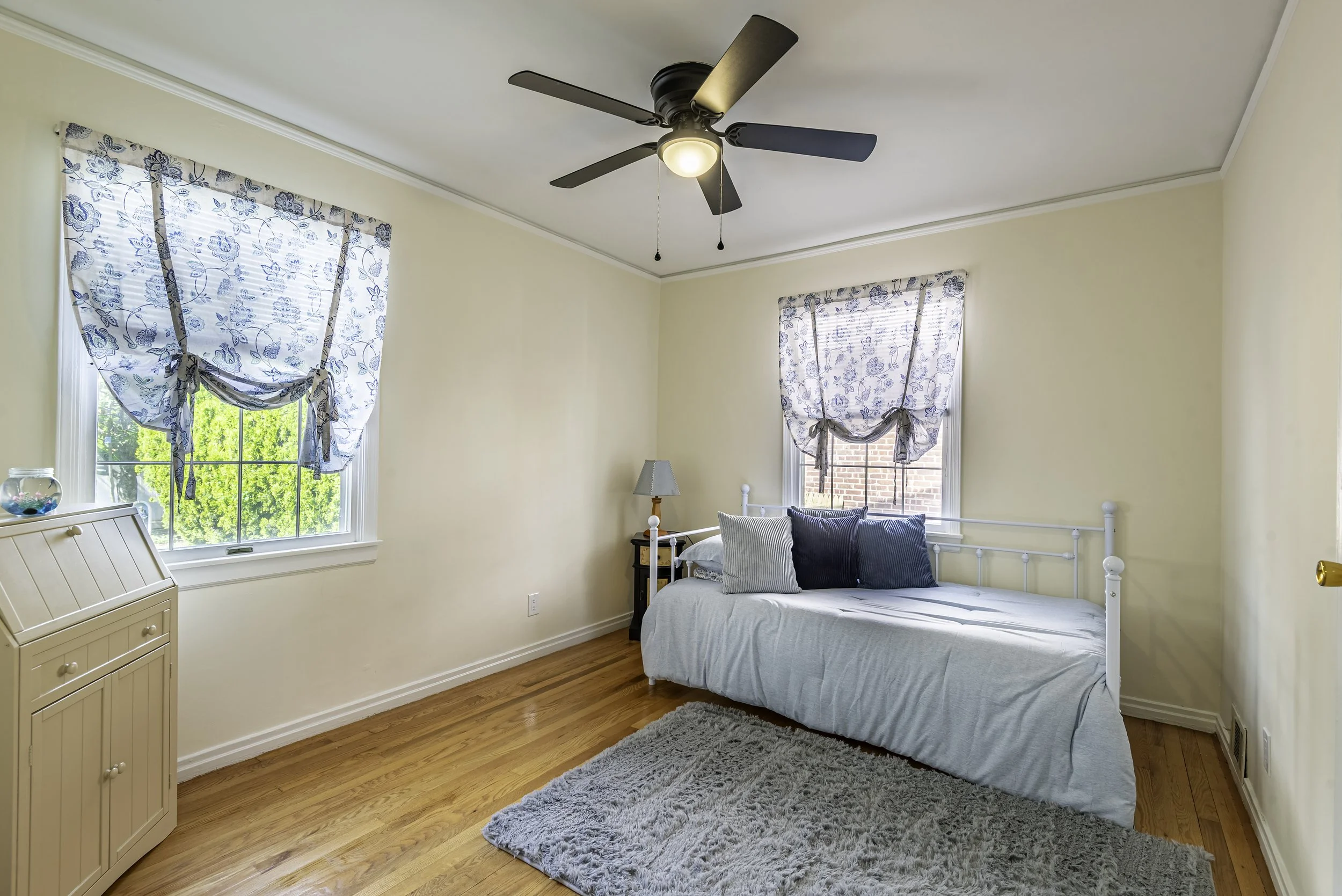 Bright bedroom with hardwood floors, ceiling fan, natural light from two windows, and cozy staged décor with daybed and area rug.