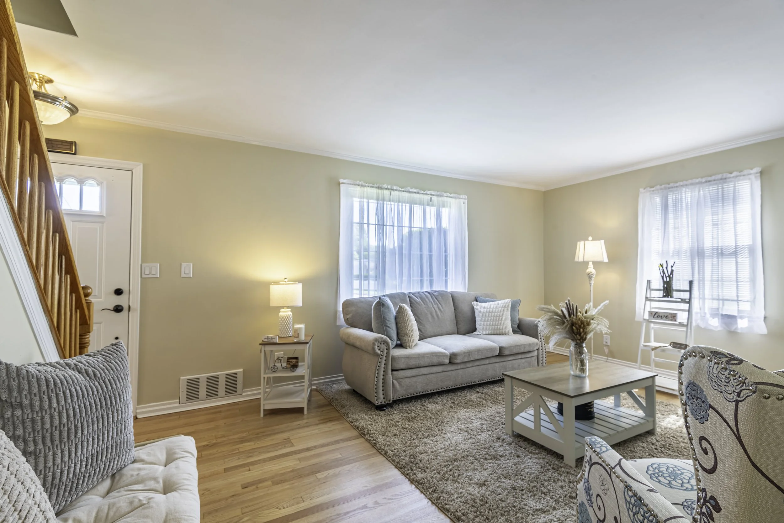 Bright and inviting living room featuring large windows, light beige walls, a gray sofa, accent chairs, and a rustic coffee table.