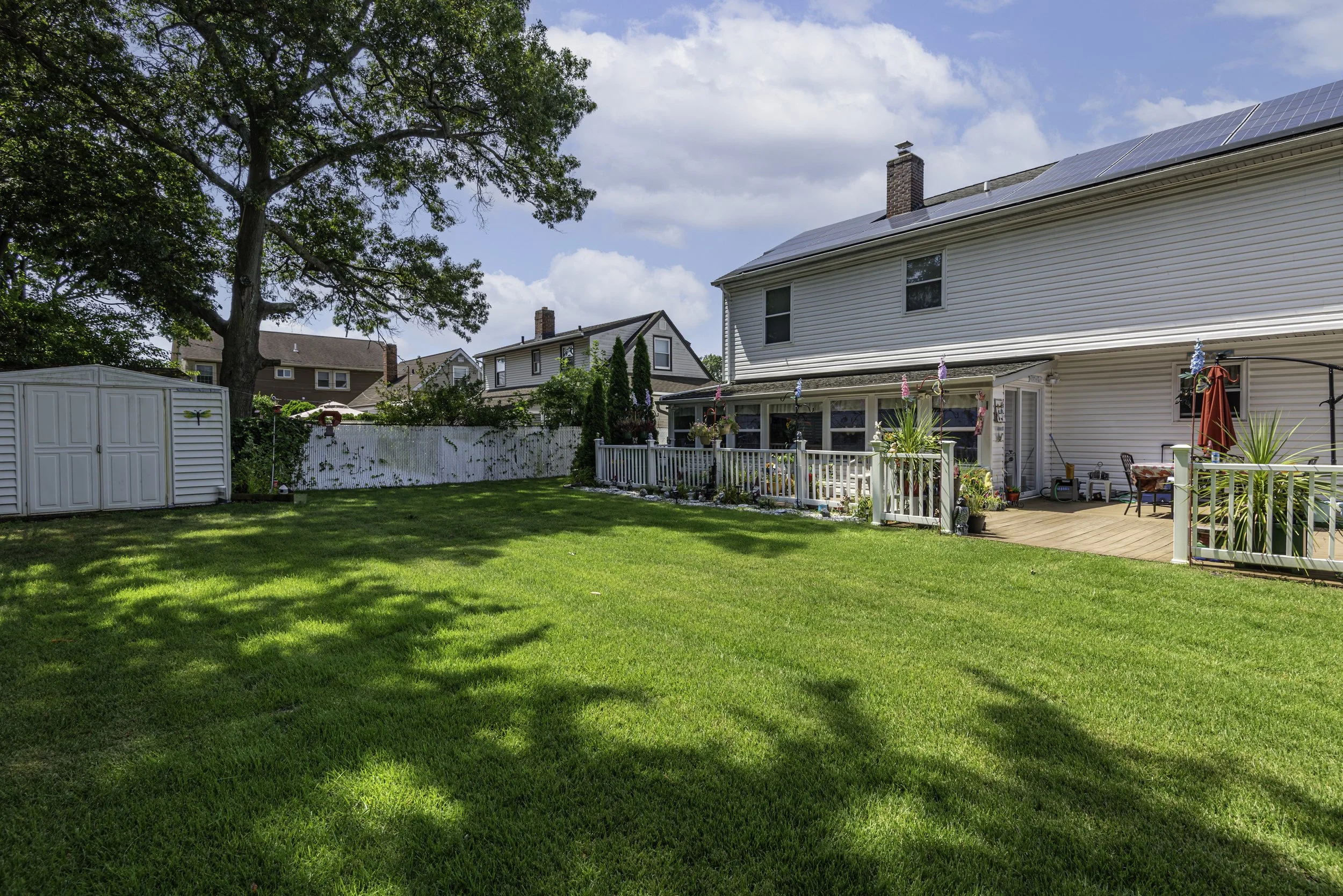 Large fenced backyard with lush green lawn, deck, storage shed, and mature trees providing shade.